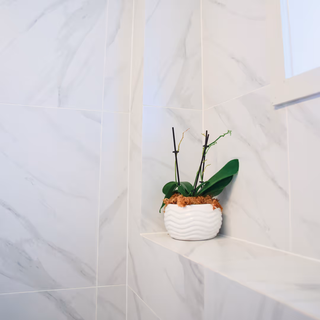 White ceramic pot with green orchid plant leaves and support stakes on marble-tiled bathroom corner shelf in modern bathroom.