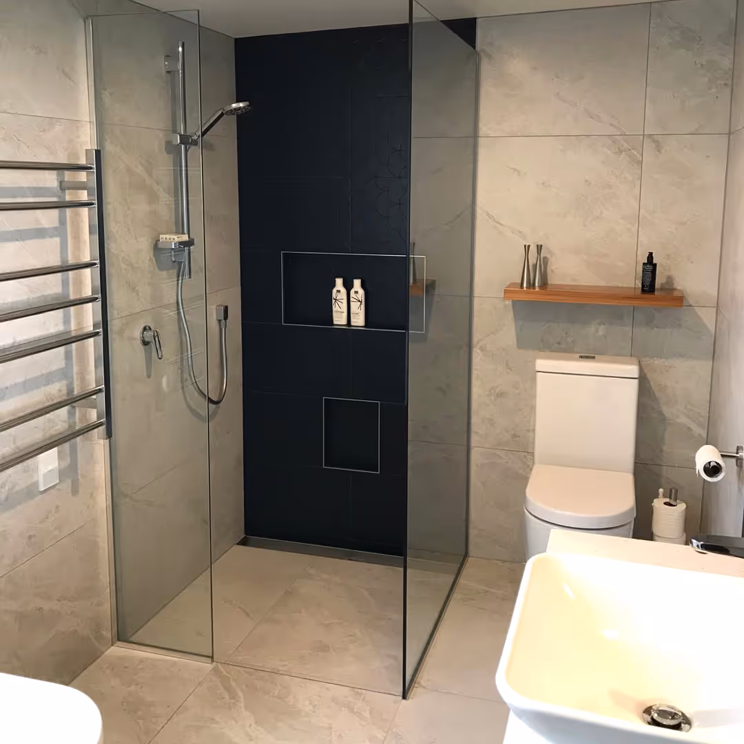Modern bathroom with glass shower enclosure, black tiled shower wall, toilet, wooden shelf, and white sink.