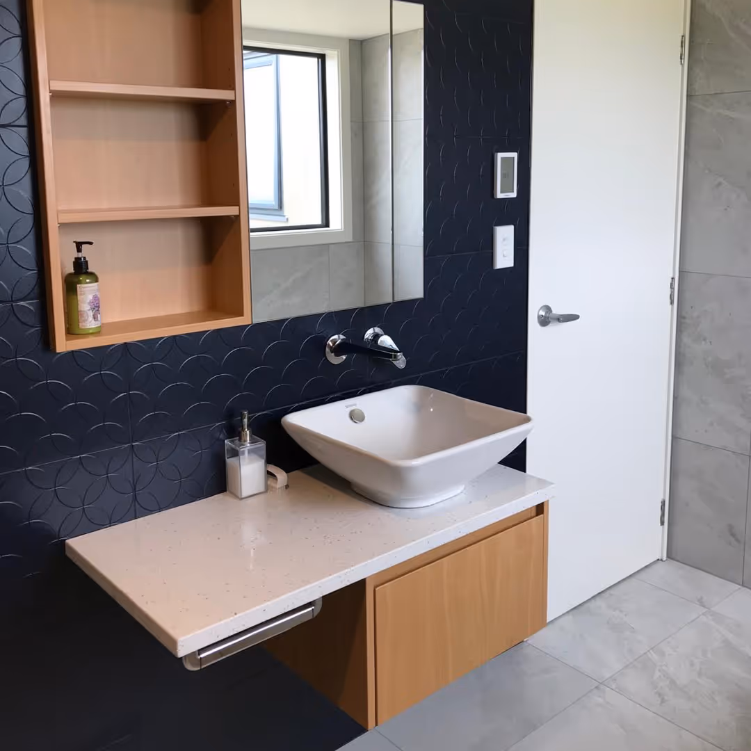 Bathroom sink with white rectangular basin, wooden cabinet, dark geometric wall tiles, and a mirror.