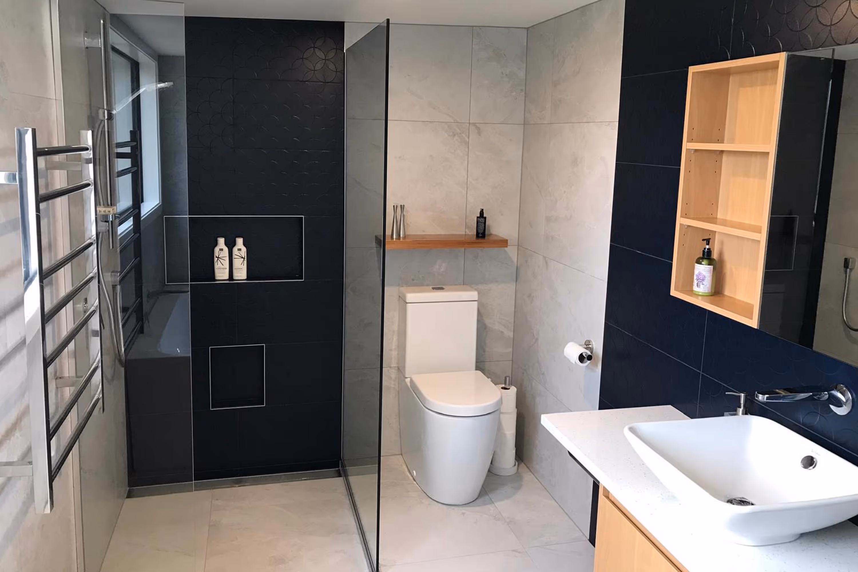 Modern bathroom with white toilet, rectangular sink, black and gray tiles, glass shower screen, and wooden shelf.