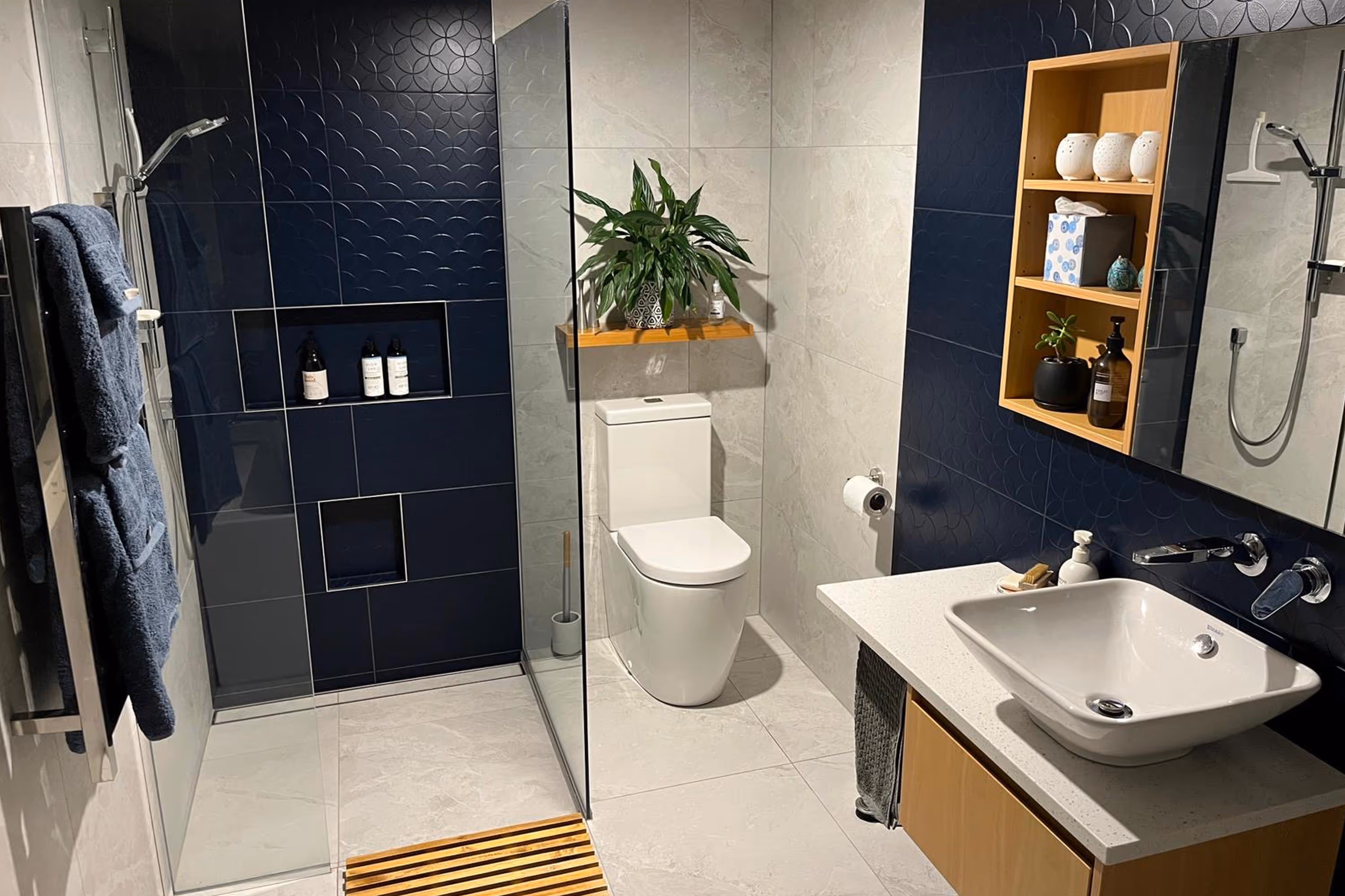Modern bathroom with navy and beige tiles, a glass shower, white toilet, sink, and wooden shelves with decor.
