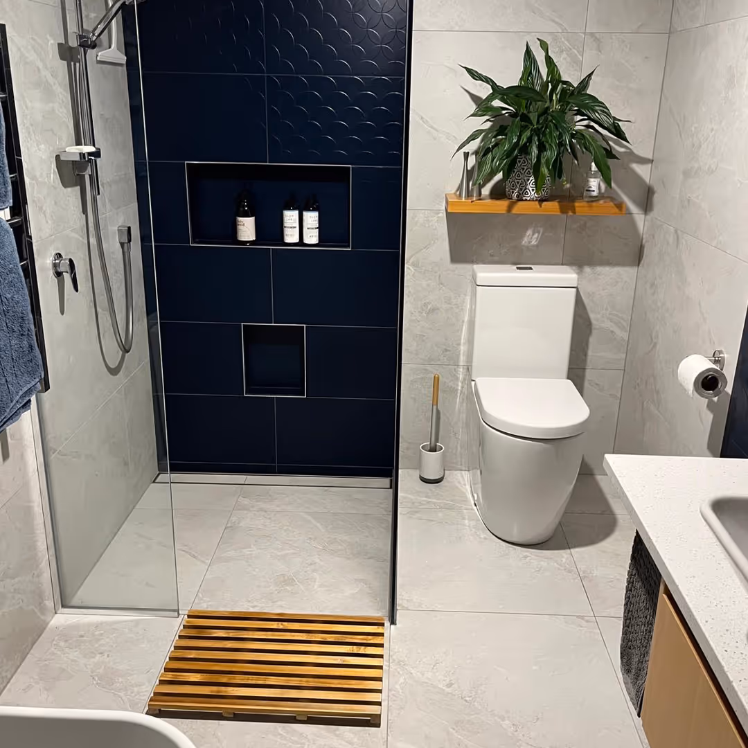 Modern bathroom with glass shower, navy tile niche, white toilet, wooden mat, and plant on shelf.
