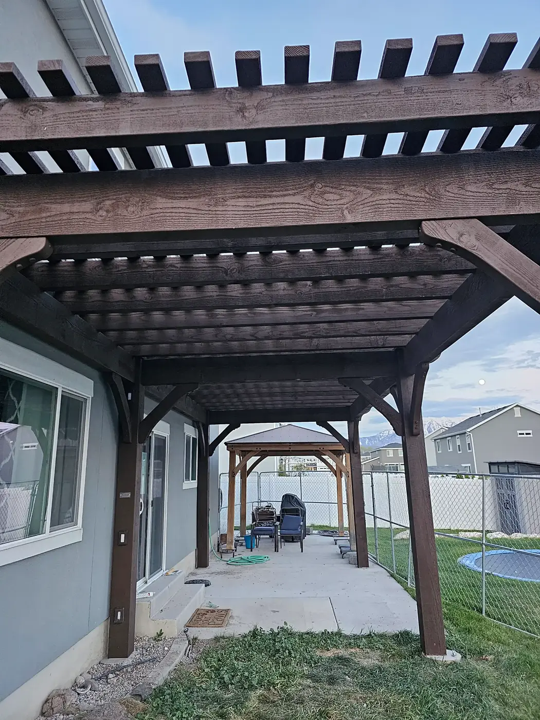 Dark-stained timber pergola with a slatted roof seen from underneath, with a second shade structure beyond