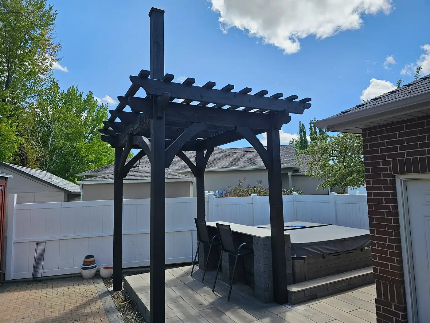 Black pergola shading a hot tub and patio seating area next to a brick wall
