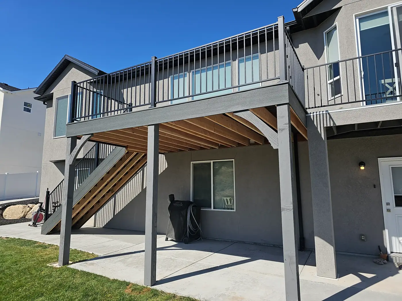 Elevated gray deck with black railing and stained joists above a concrete patio