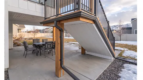 Drainage trough and downspout under an elevated deck stair above a dry concrete patio