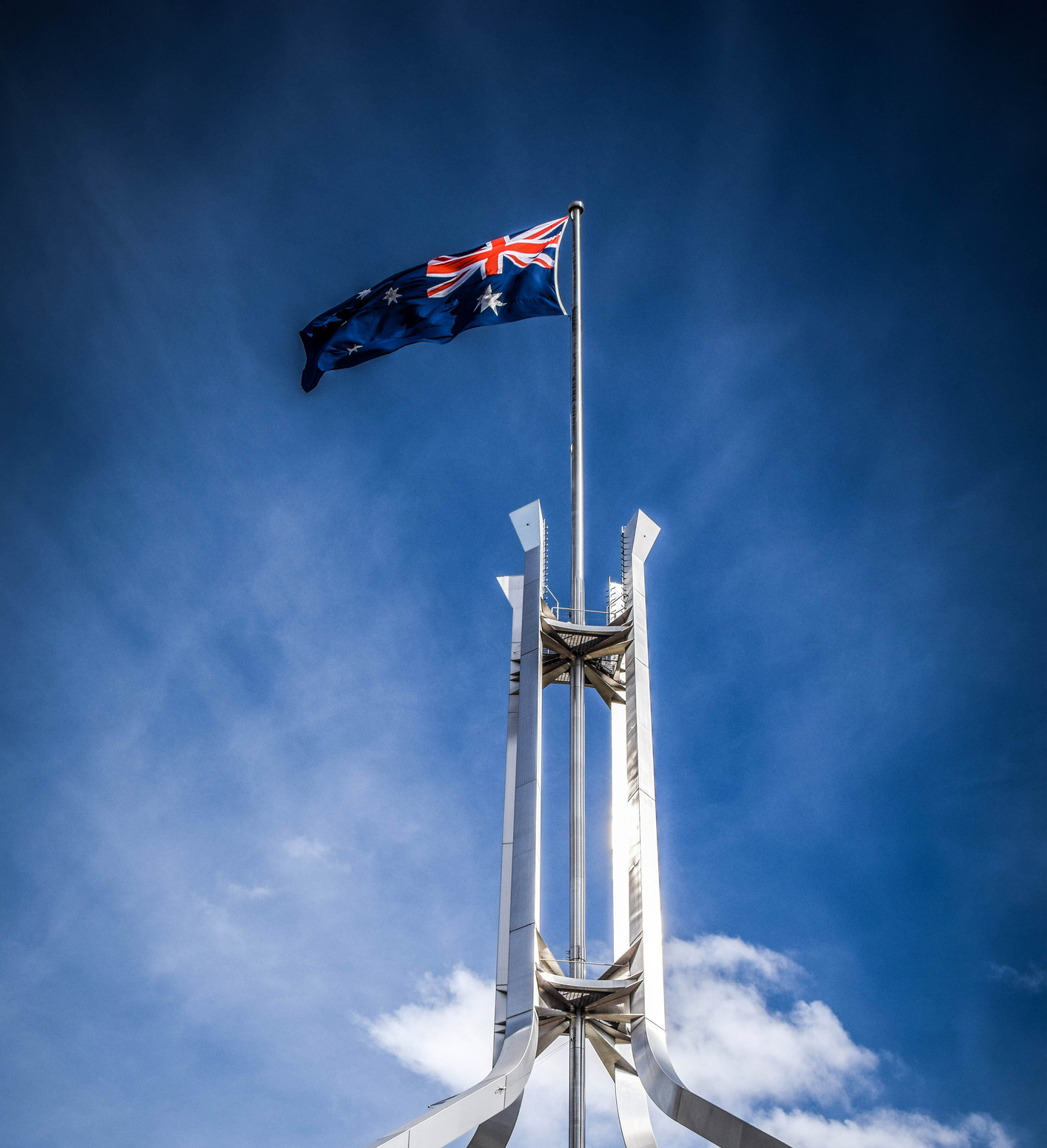 Australian flag flying atop the metallic structure of the Parliament House flagpole against a clear blue sky.