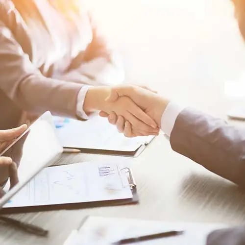 Two business professionals in white shirts shaking hands over documents at a table.