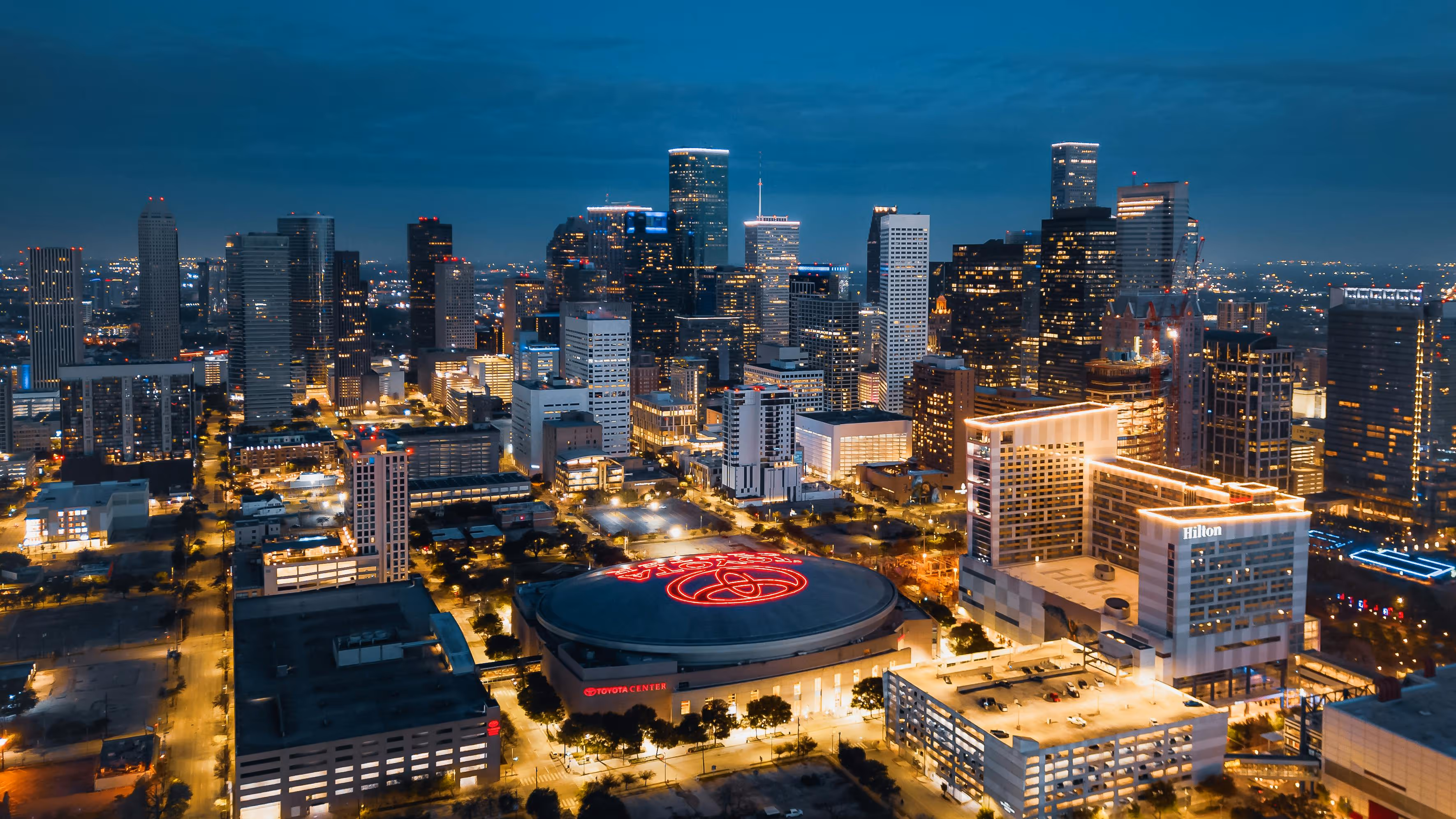 Night aerial view of Houston skyline with illuminated Toyota Center and Hilton hotel.
