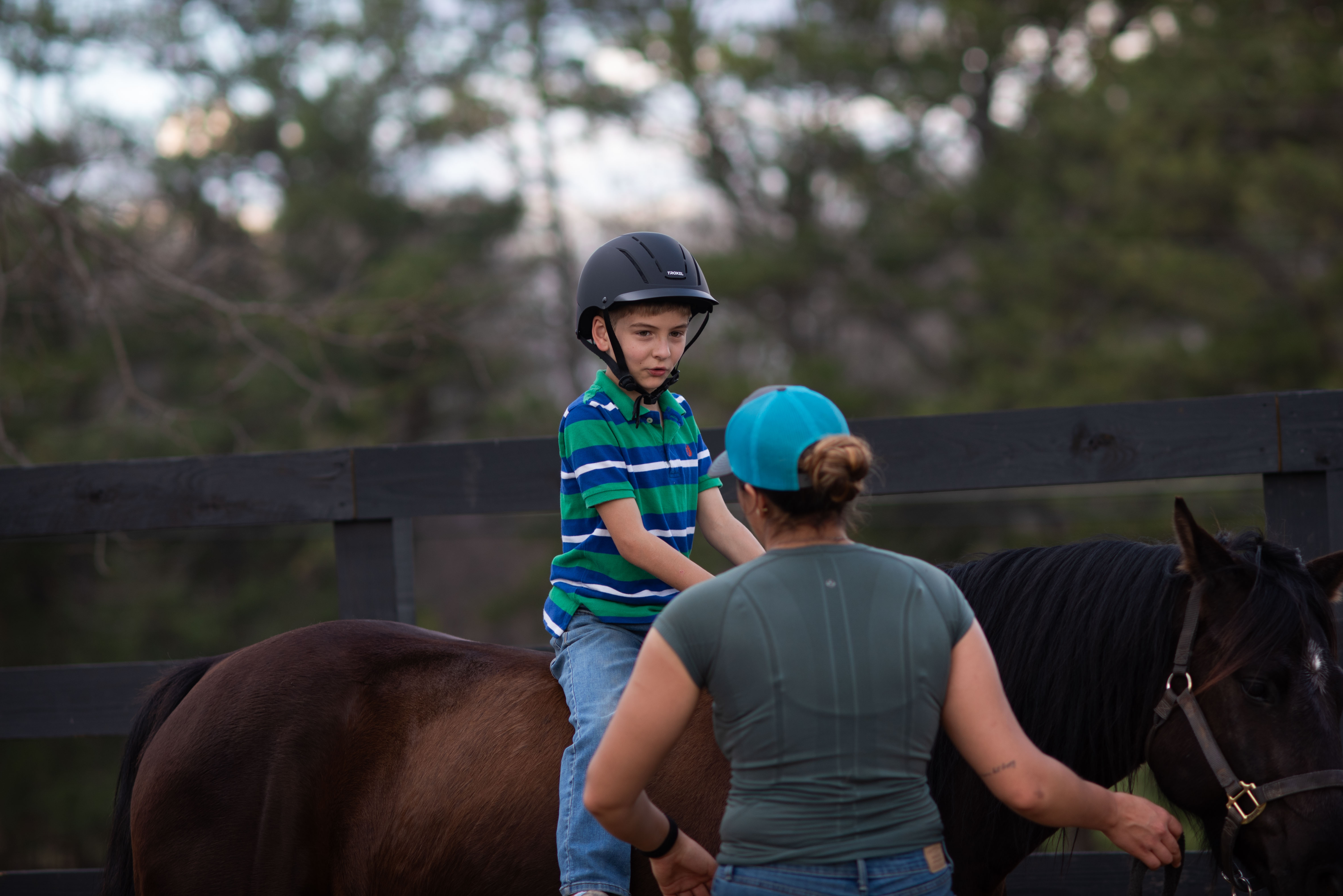 Young boy wearing a helmet sitting on a brown horse while an adult woman with a blue cap stands in front of the horse.