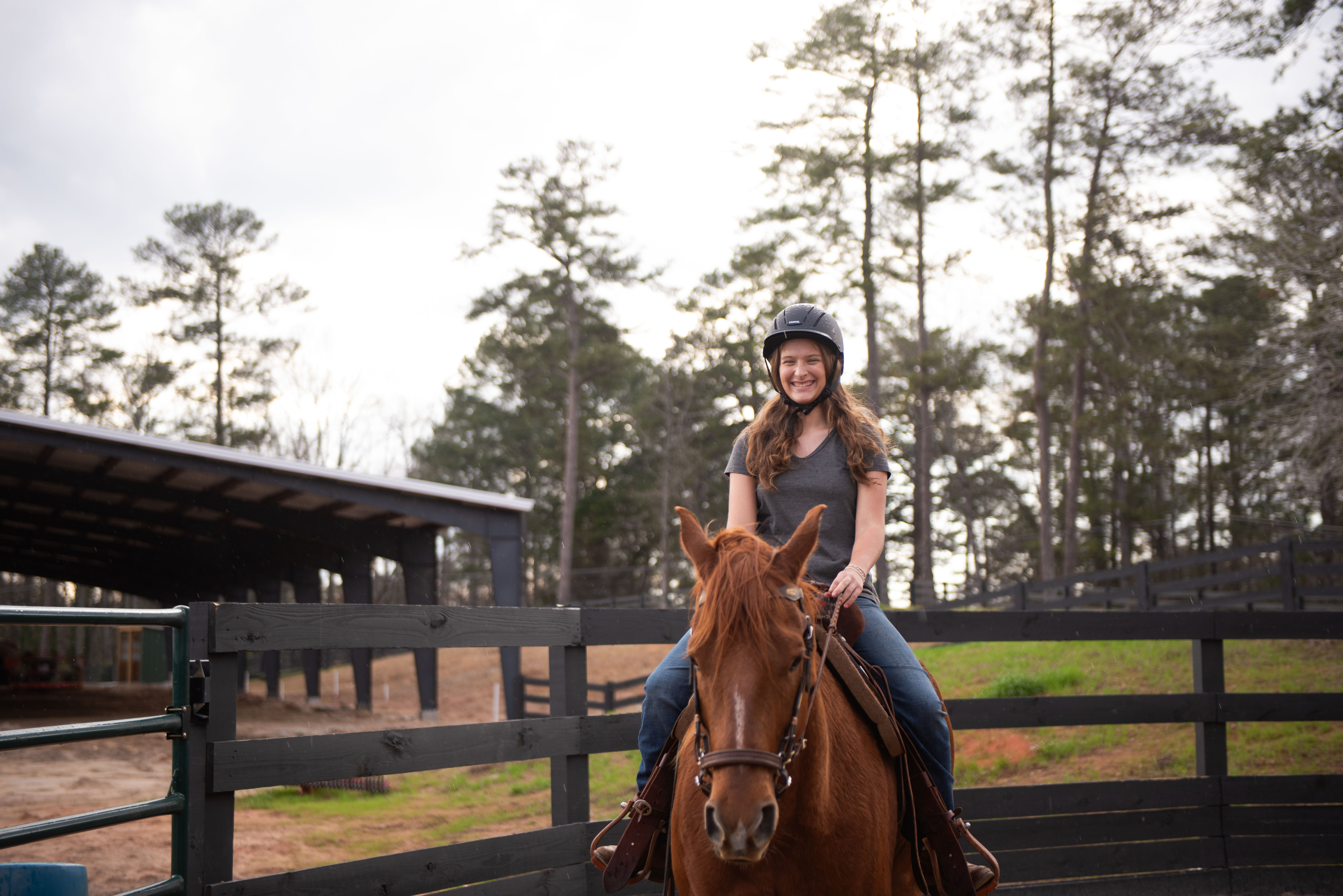 Teenage girl wearing a helmet riding a brown horse near a fenced riding area with trees in the background.