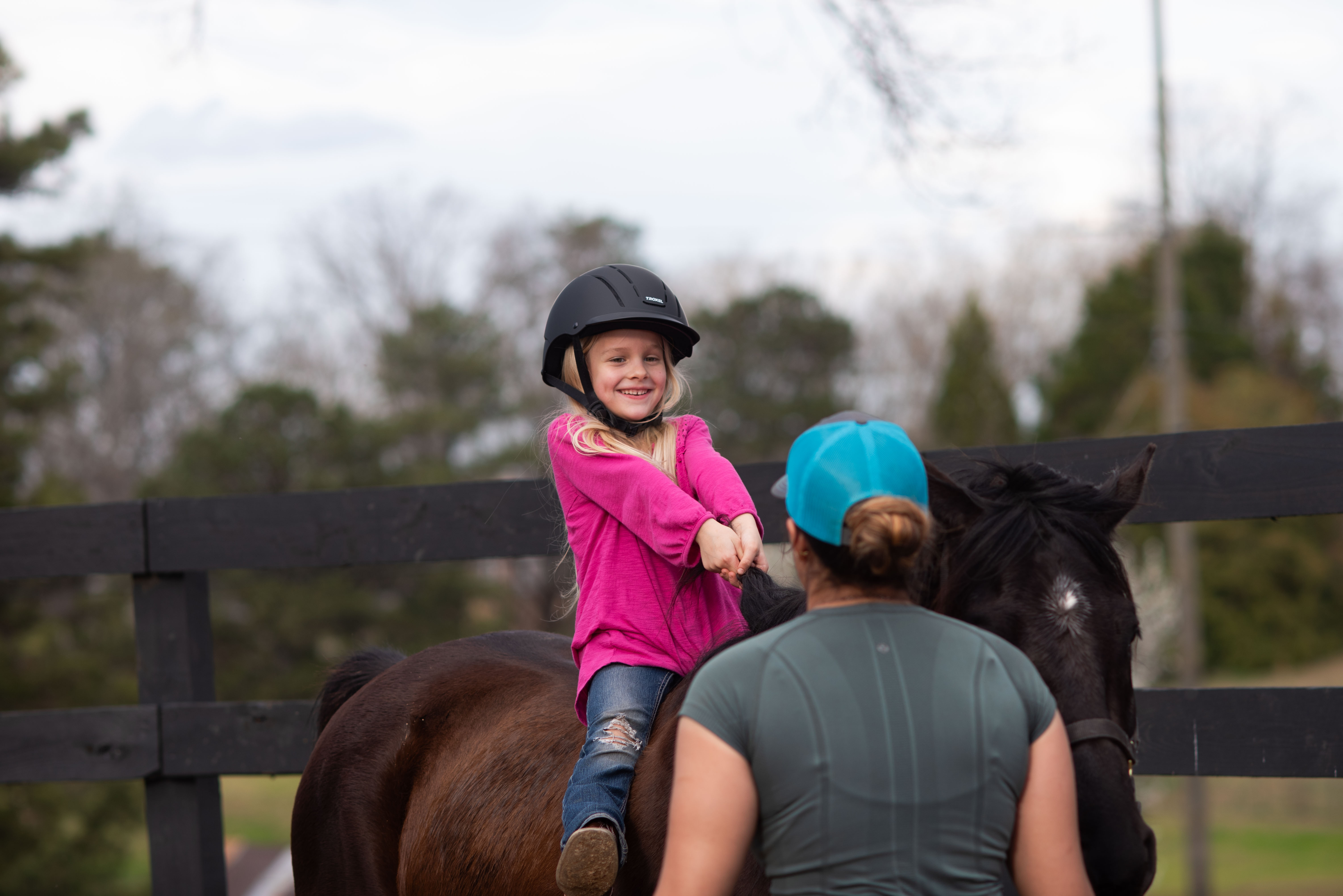 Young girl in a helmet and pink shirt smiling while sitting on a dark horse, with an adult wearing a blue cap standing beside the horse.