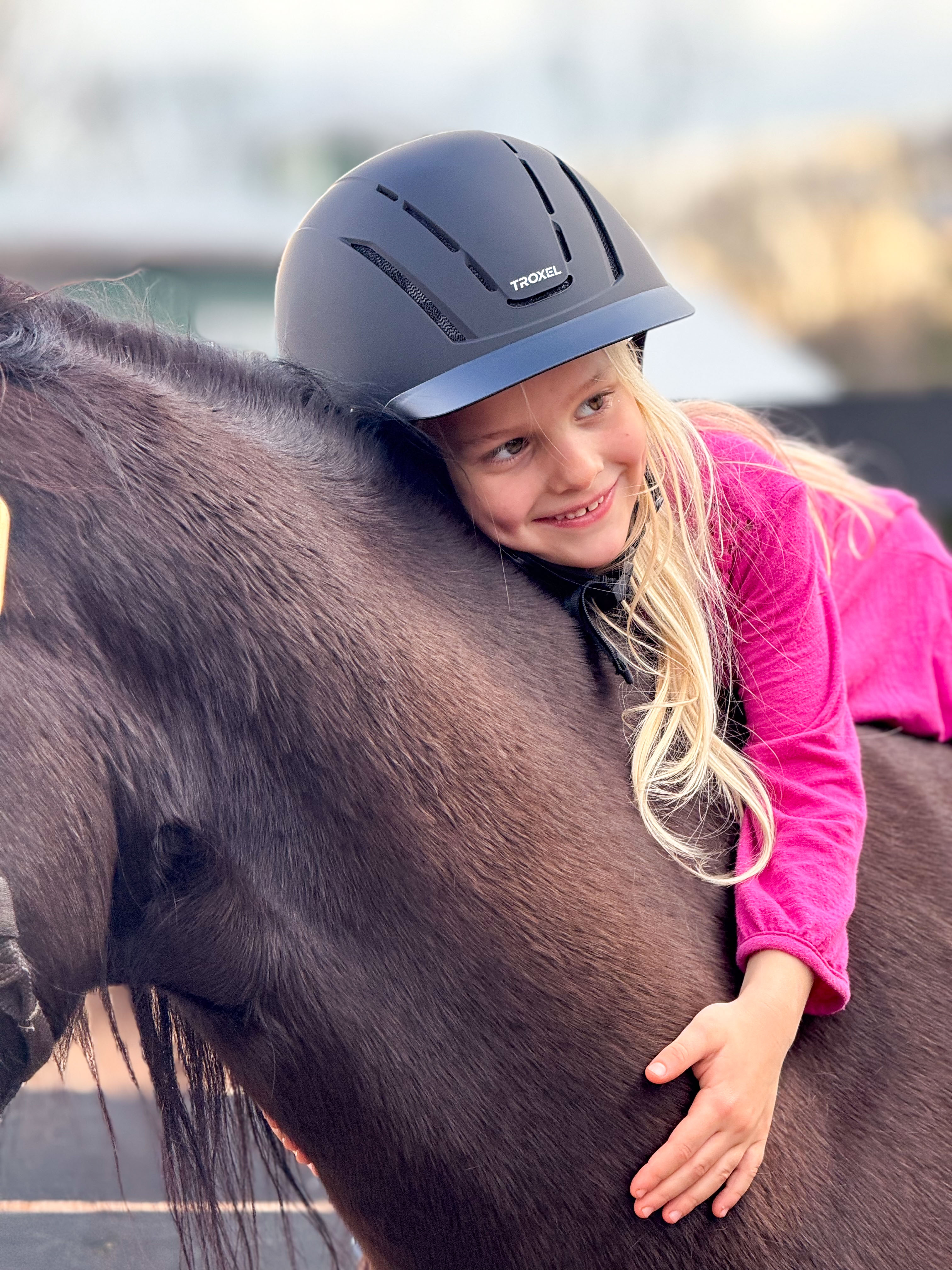 Smiling girl in a black riding helmet and pink jacket hugging a dark brown horse.