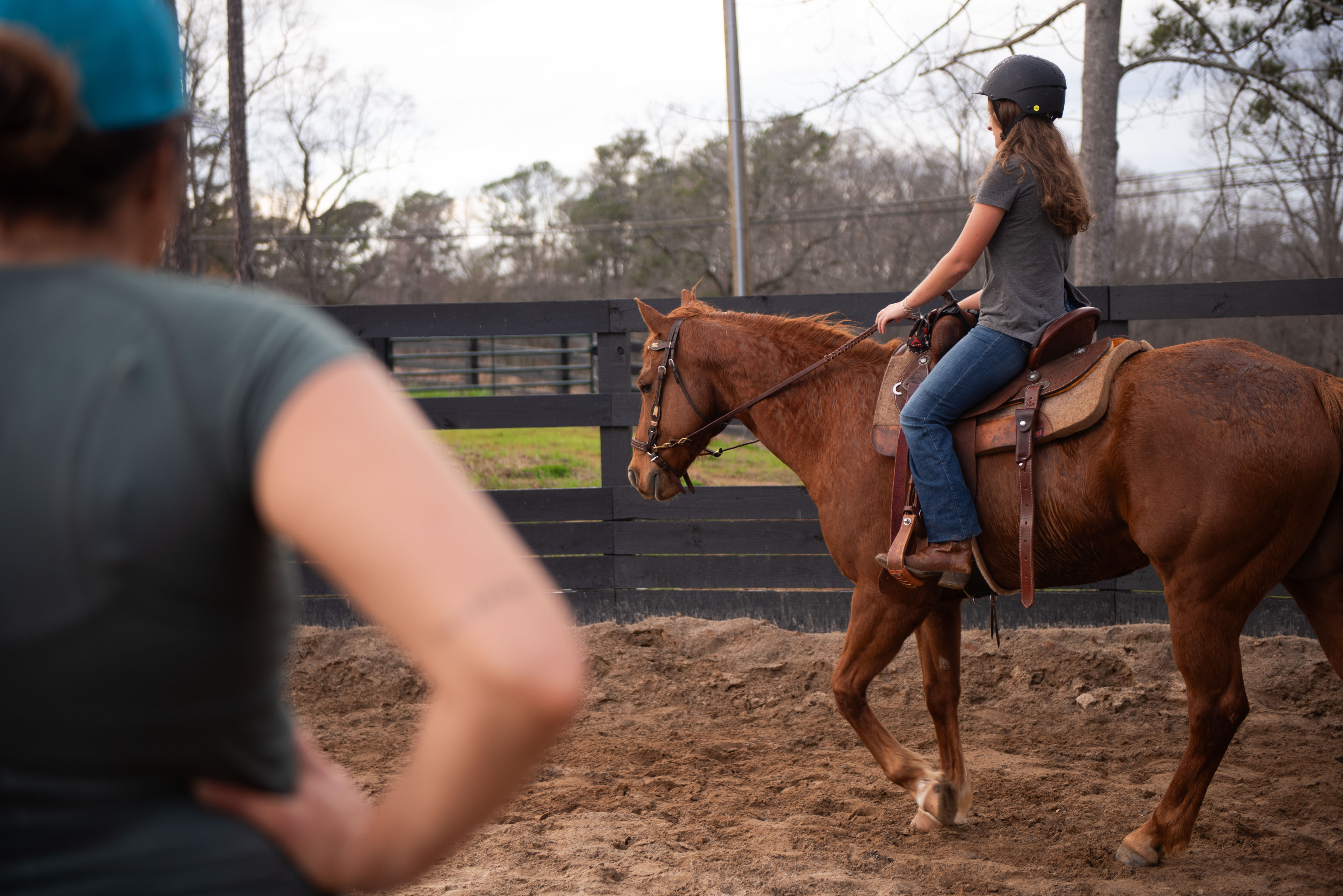 Young girl wearing helmet riding a brown horse in an outdoor arena with a person observing in the foreground.