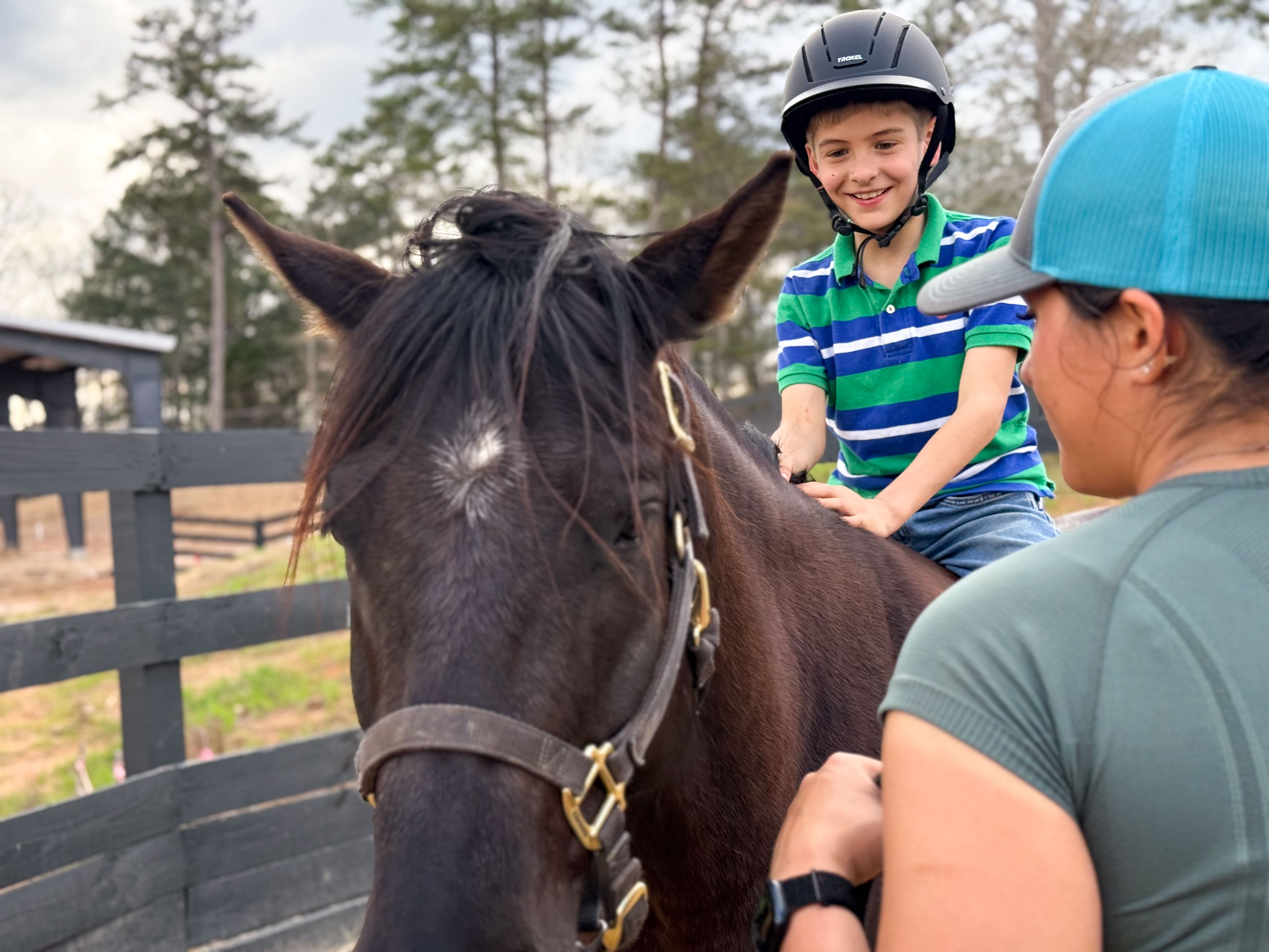 Boy wearing a helmet and striped green and blue shirt smiling while sitting on a black horse, assisted by a woman in a teal cap and shirt.