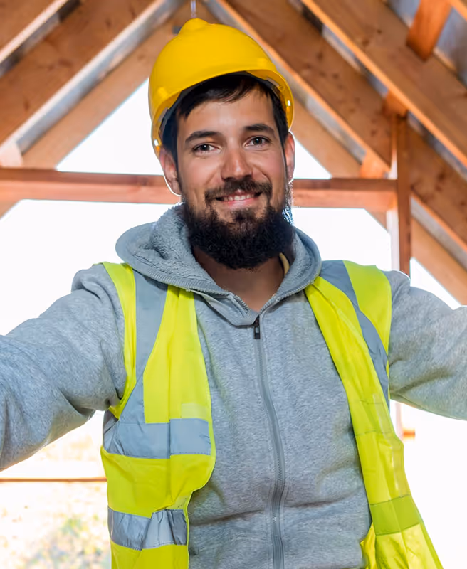 Smiling male construction worker wearing a yellow hard hat and high-visibility vest indoors with wooden beams in the background.