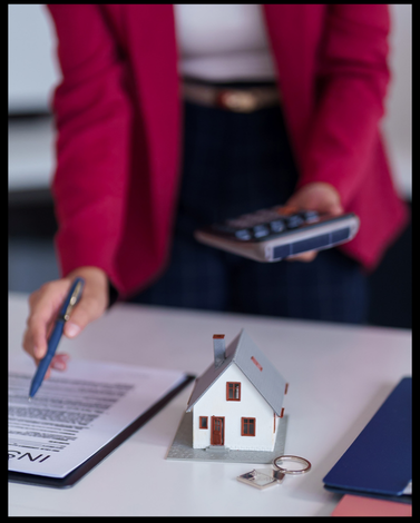 Person holding a pen and calculator near a small model house, contract, and house keys on a table.