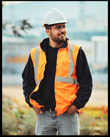 Smiling male construction worker wearing a white helmet, glasses, black shirt, and an orange safety vest with hands in pockets.