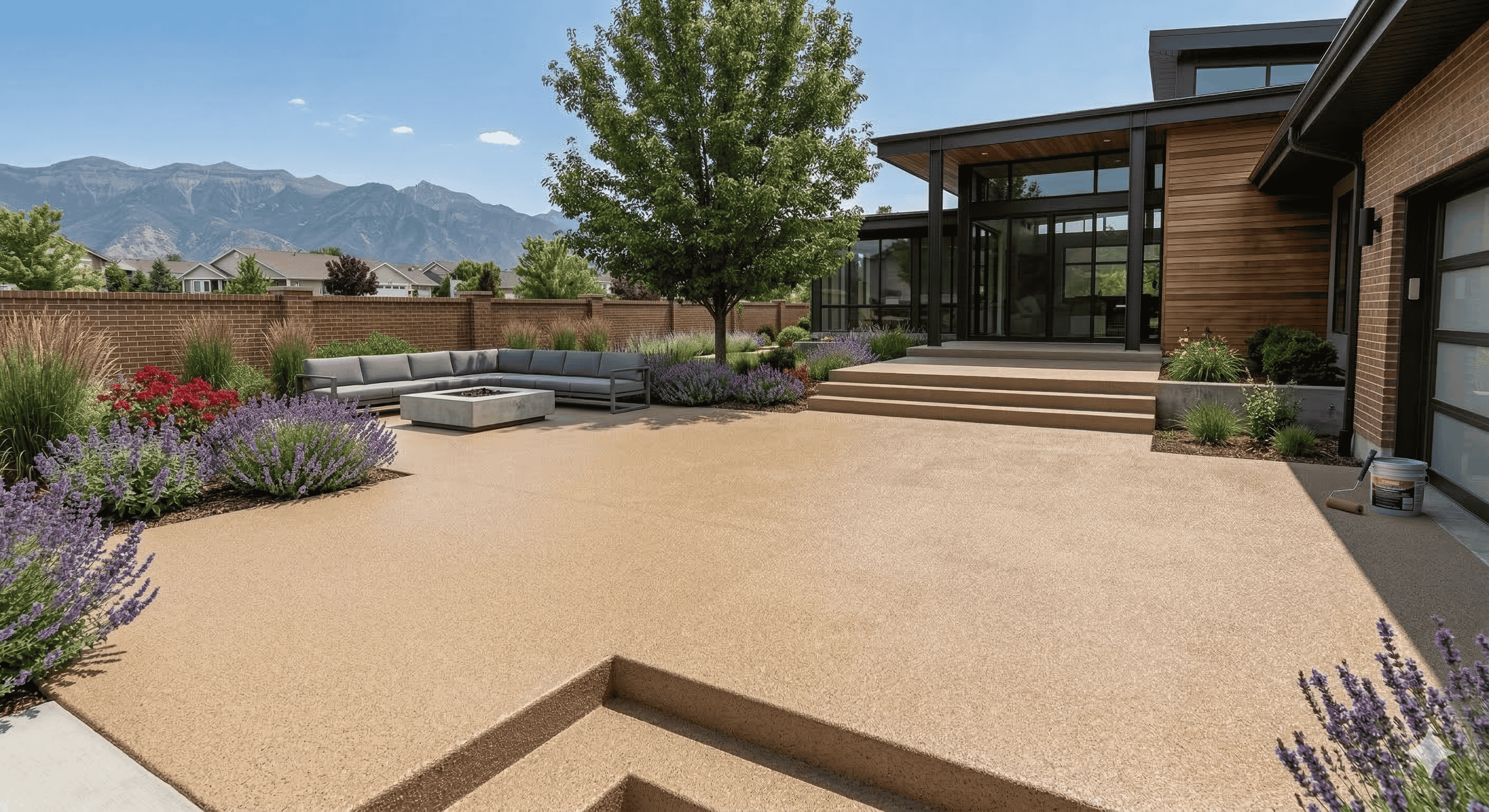 Modern backyard patio with beige textured flooring, gray sectional sofa around a fire pit, and mountain view behind a brick wall.