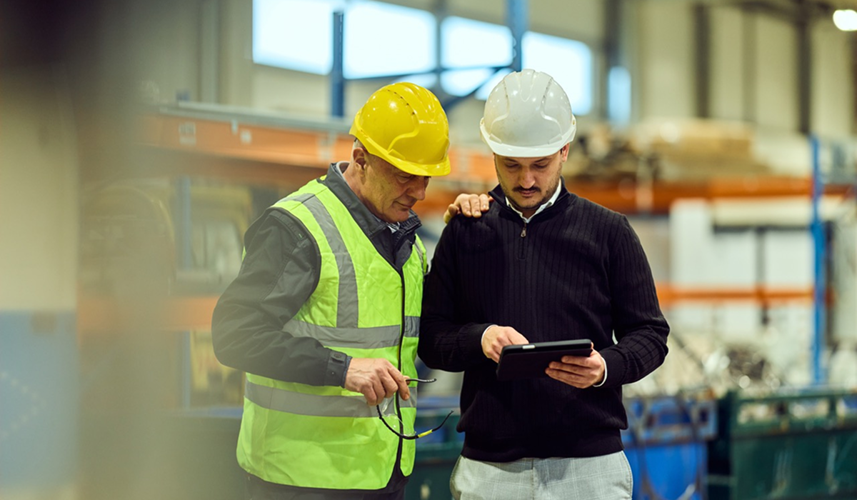 Two people wearing hard hats review a tablet together in an industrial workspace.