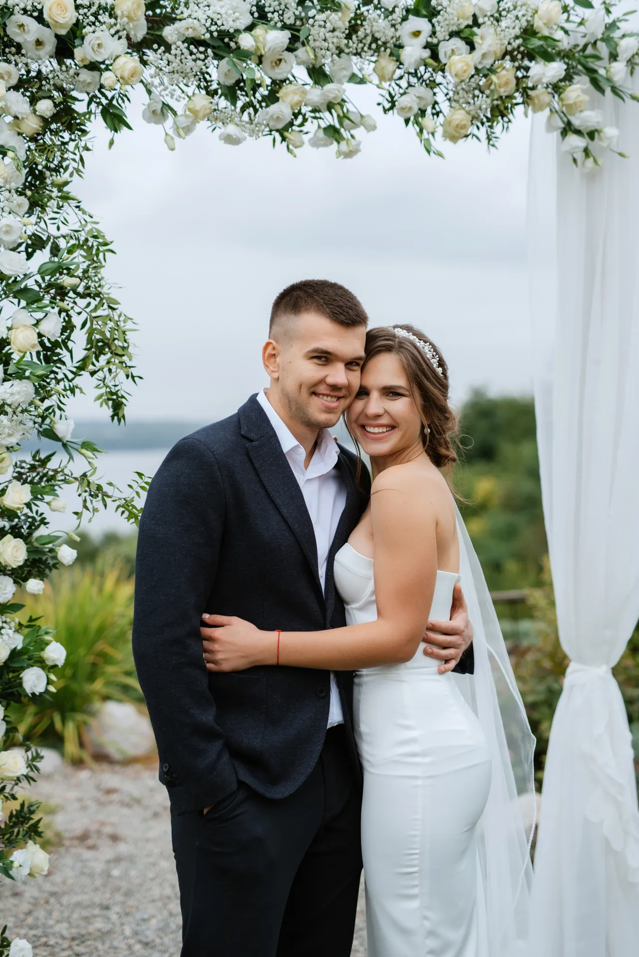 Brudepar omfavner hinanden under en blomsterbue med hvide roser og grønne blade udendørs.