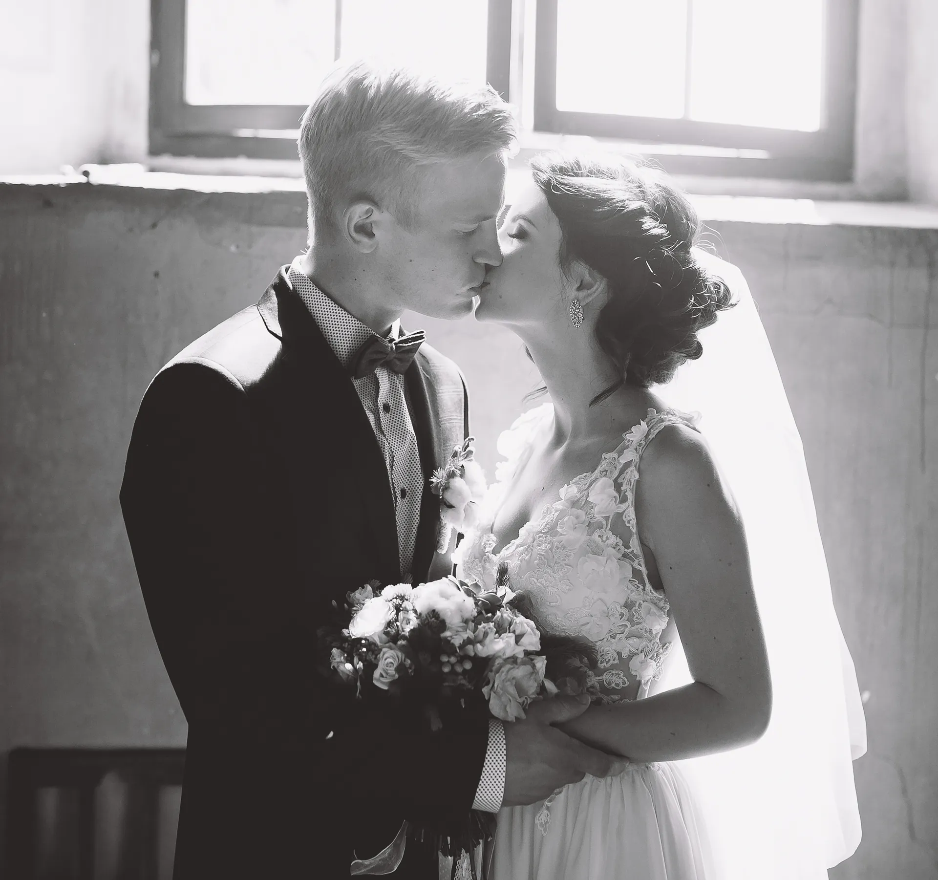 Brud og gom kysser foran et oplyst vindue, bruden holder en blomsterbuket.