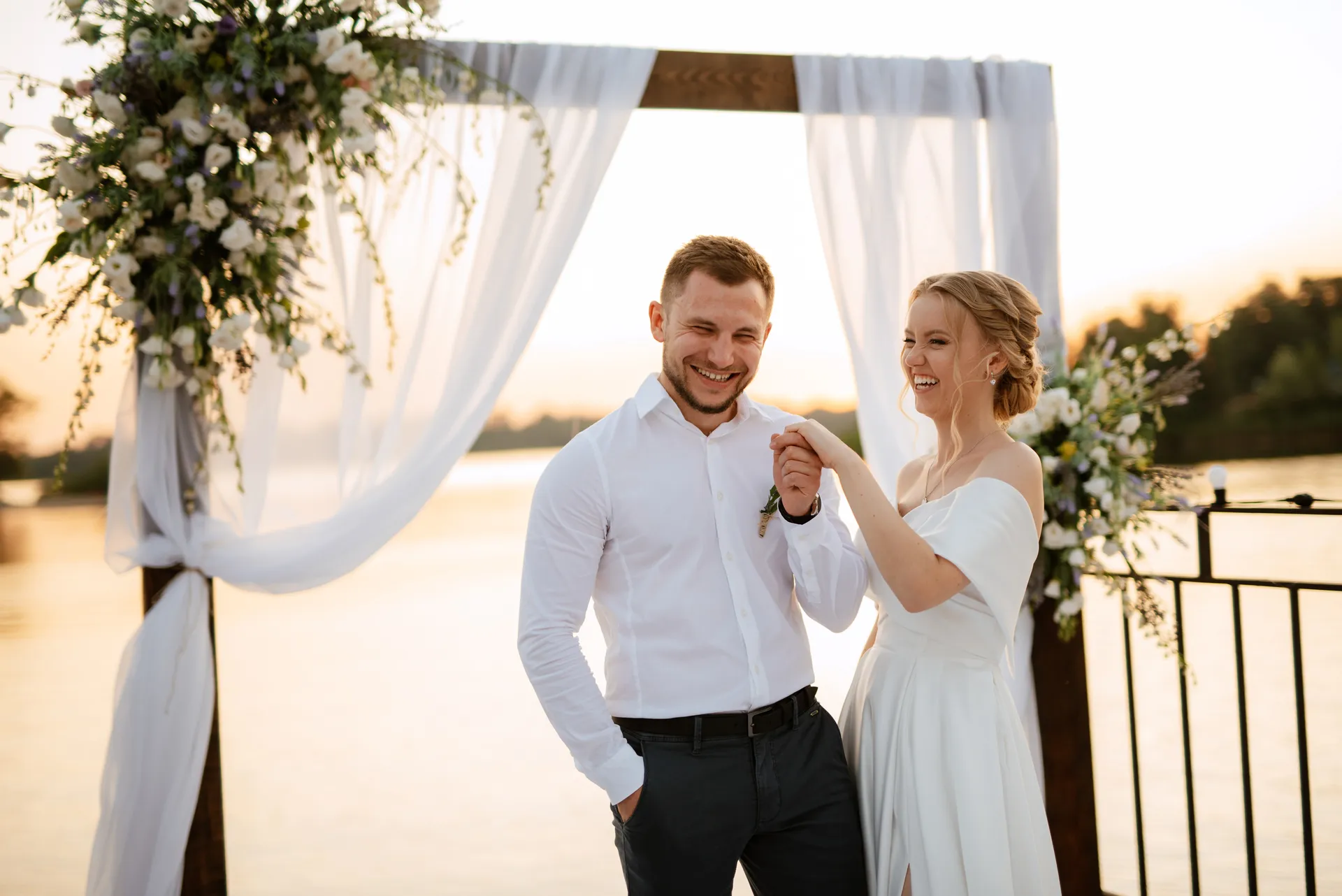 Brud og gom holder hinandens hænder og ler ved en dekoreret bænk med udsigt over vandet ved solnedgang.