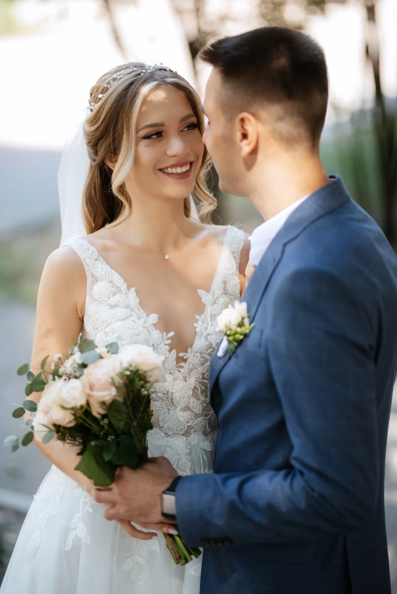 Brudepar med brudebuket, hvor bruden smiler til gommen iført en blondebrudekjole og gommen en blå jakkesæt.
