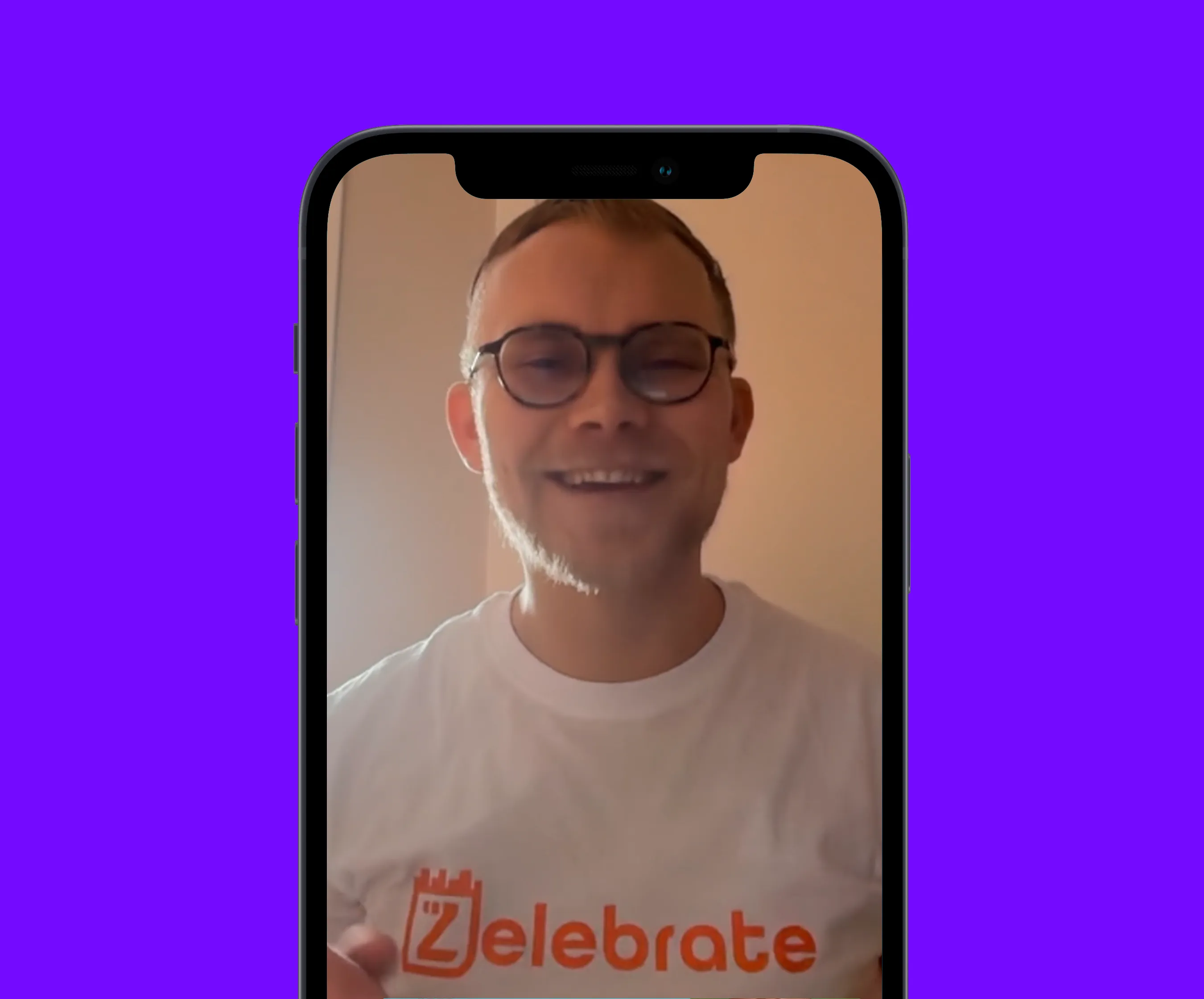 Mand iført briller og en hvid t-shirt med teksten Zelebrate på, smilende inde i en smartphone-skærm med lilla baggrund.
