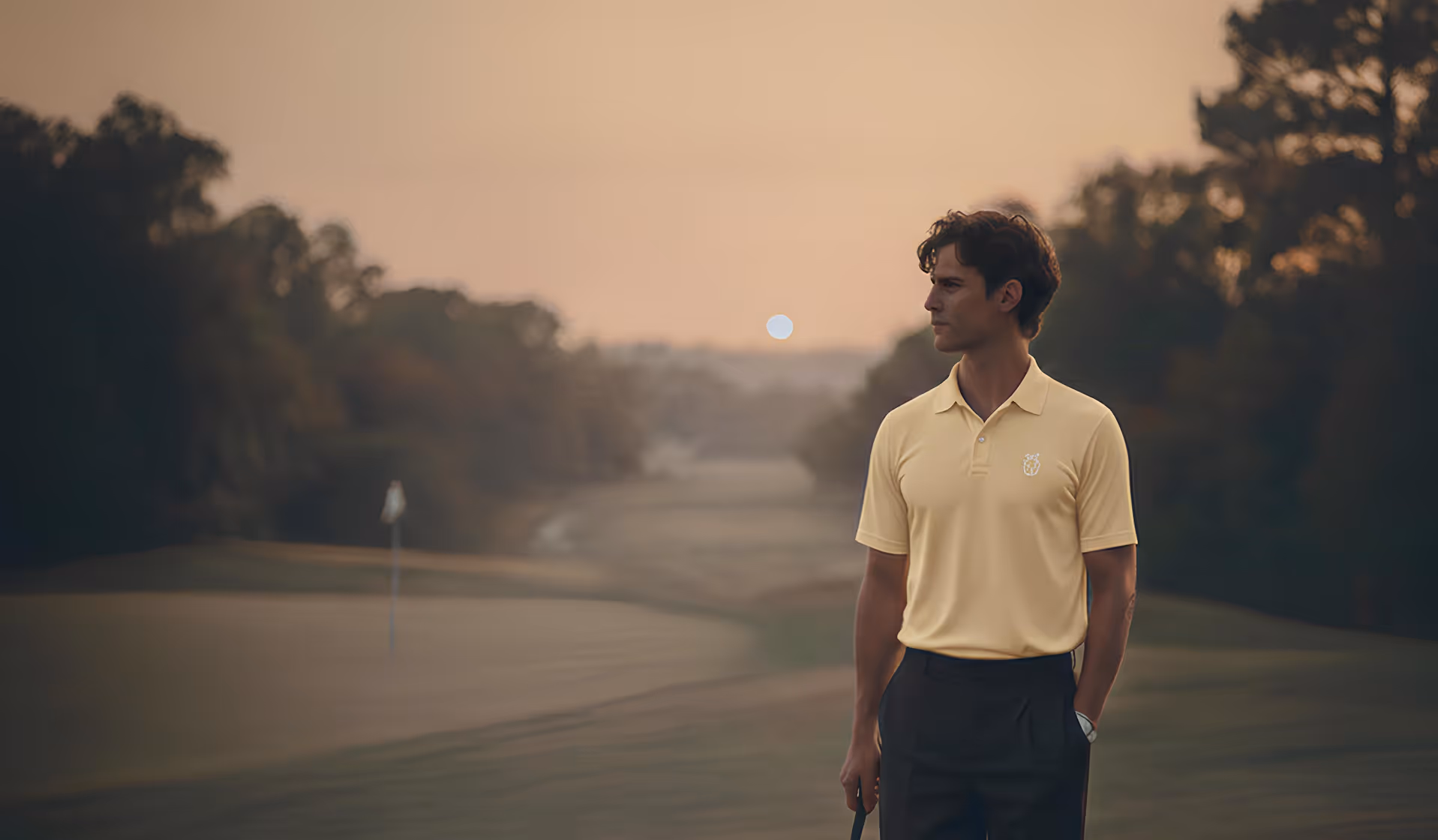 Man in yellow polo shirt standing on a golf course at sunset, holding a golf club and looking to the side.