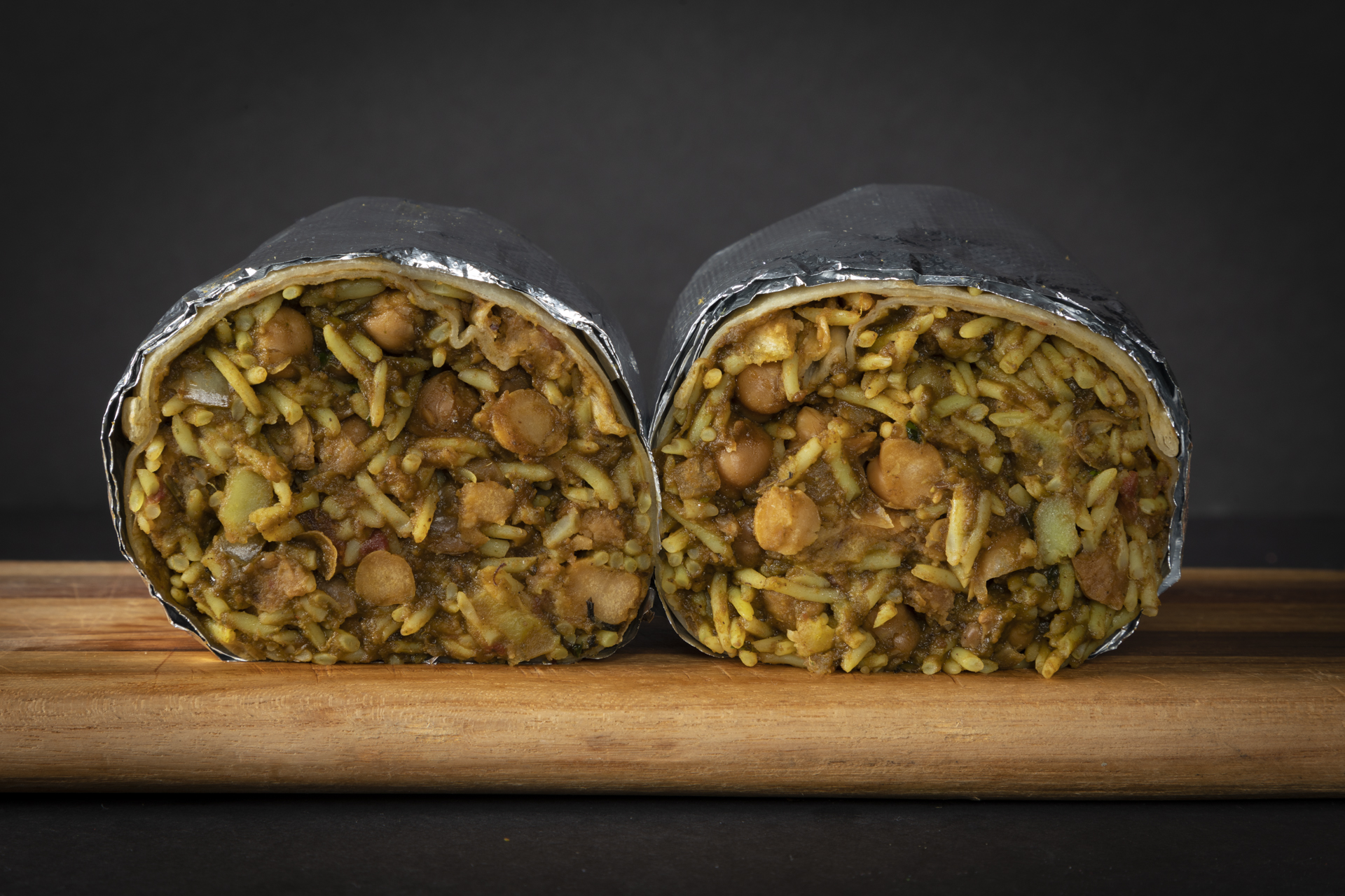 Two halves of a chickpea and rice burrito wrapped in foil on a wooden board.