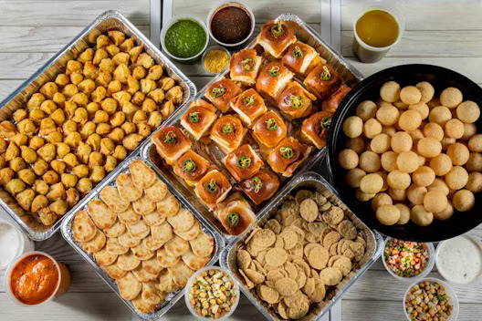 Tray of Indian street food including pani puri, samosas, pav bhaji, chutneys, and various fried snacks served with garnishes.