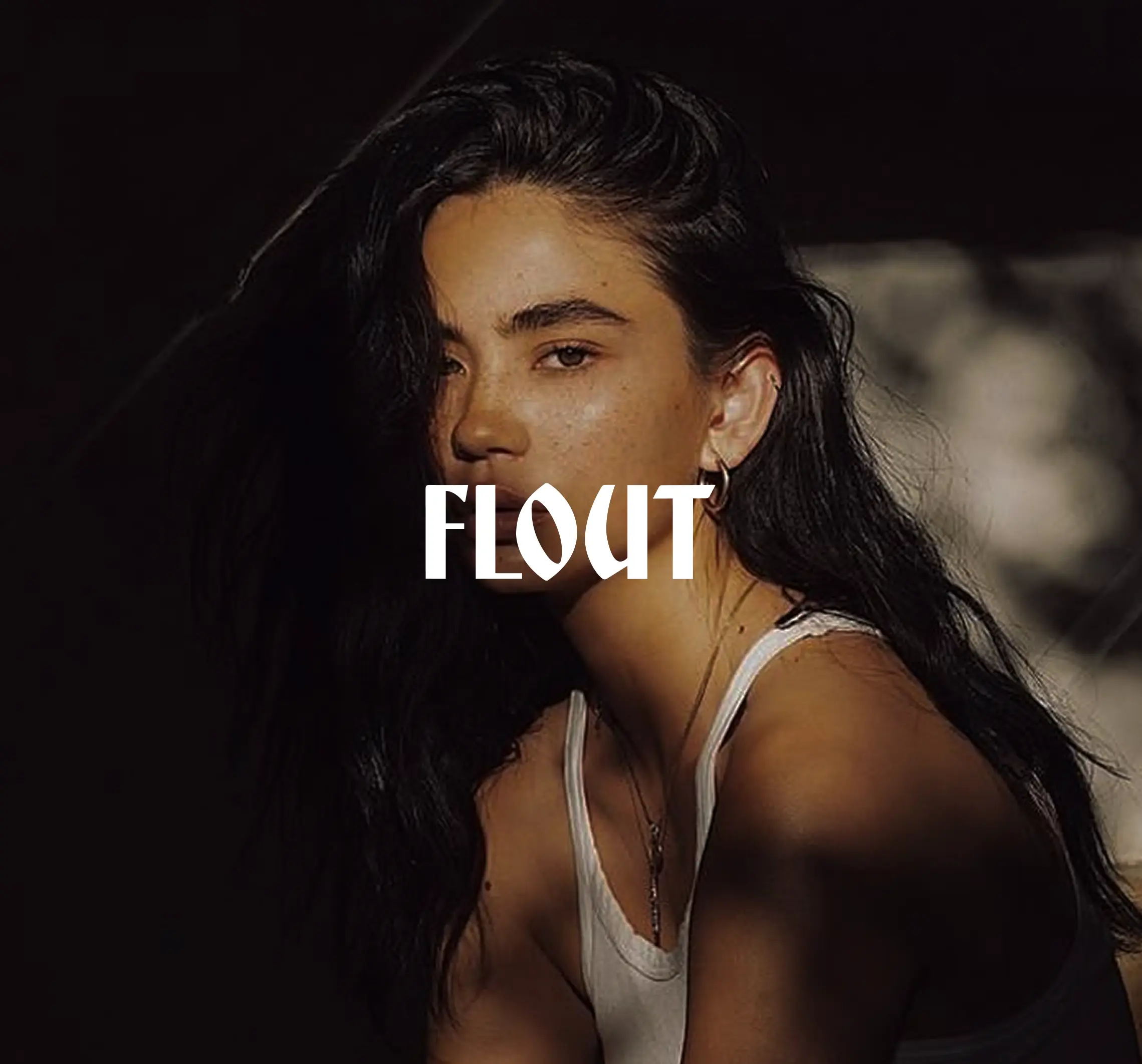 Young woman with long dark hair in a white tank top, partially illuminated against a dark background with the word 'FLOUT' overlaid.