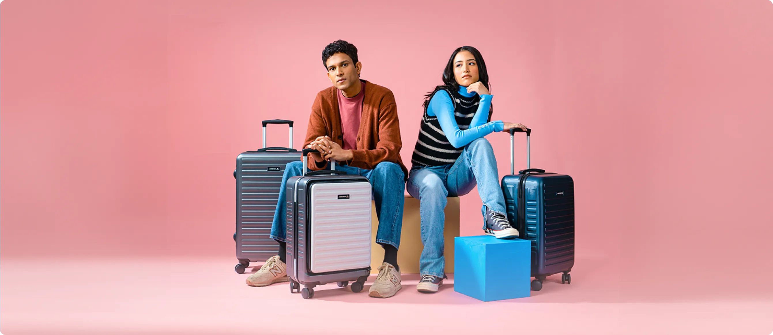Man and woman sitting with three hard-shell rolling suitcases against a pink background; man wears brown cardigan and jeans, woman wears blue long sleeve and jeans, seated on cube stools.