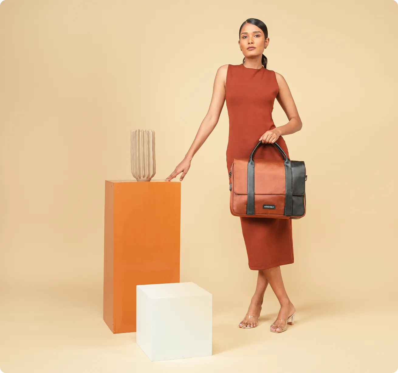 A woman in a sleeveless rust-colored dress stands next to a tall orange pedestal with a beige vase on top, holding a large brown and black leather bag with the label 'ASSEMBLY' in front of a beige background.