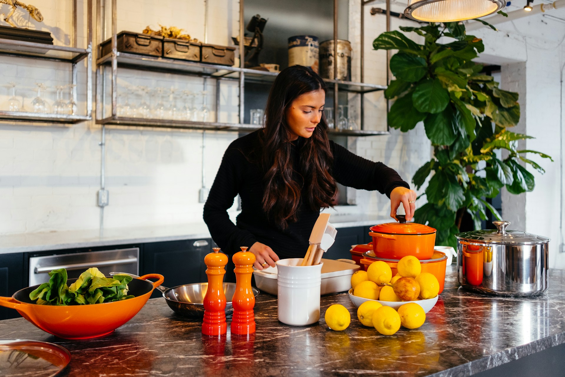 A woman prepares food in a bright, industrial-style kitchen, lifting the lid from an orange pot on a marble island surrounded by lemons, leafy greens, cookware, utensils and matching orange pepper mills.