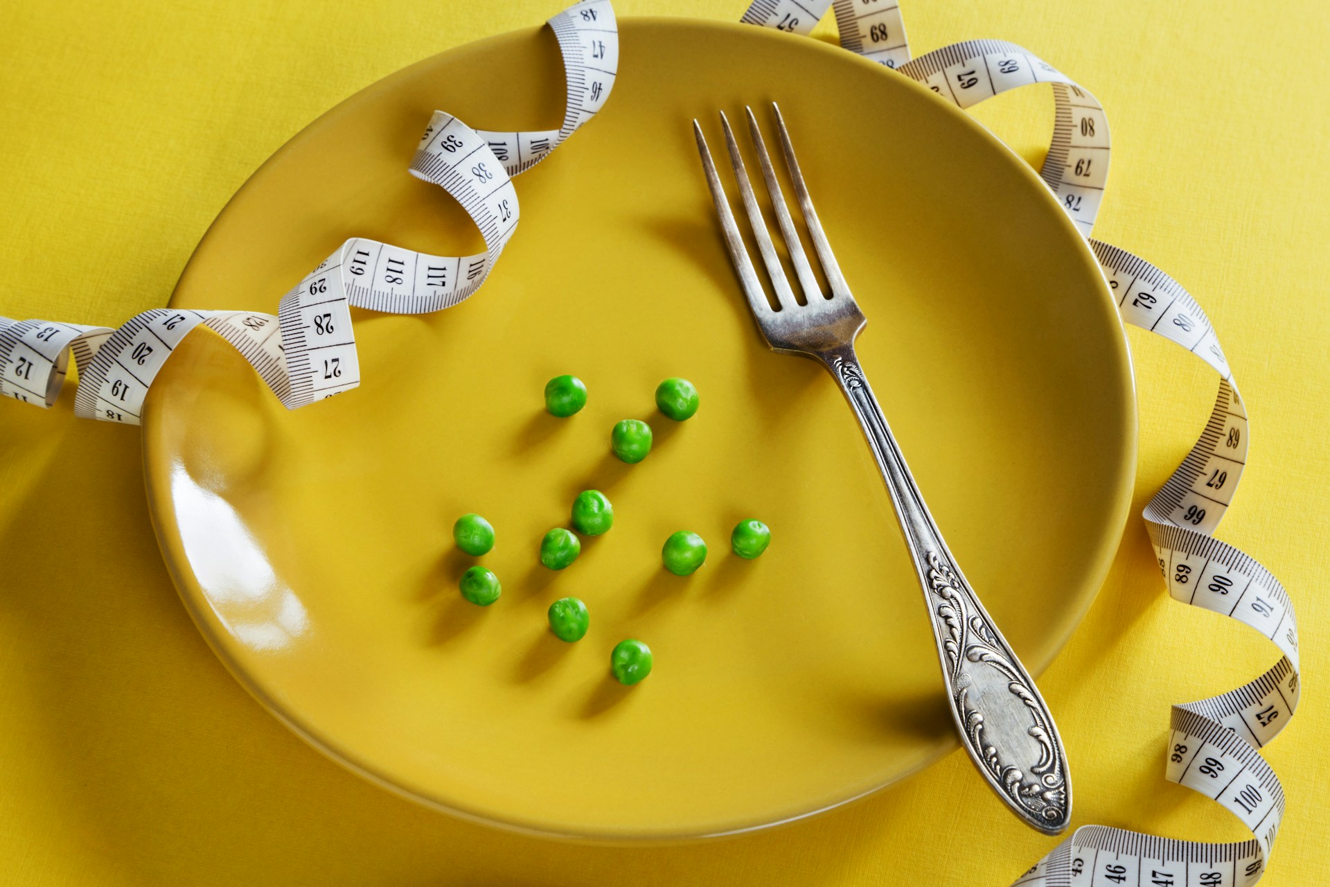 A yellow plate holds a fork and a small serving of green peas, surrounded by a curled measuring tape on a matching yellow background, suggesting dieting or portion control.
