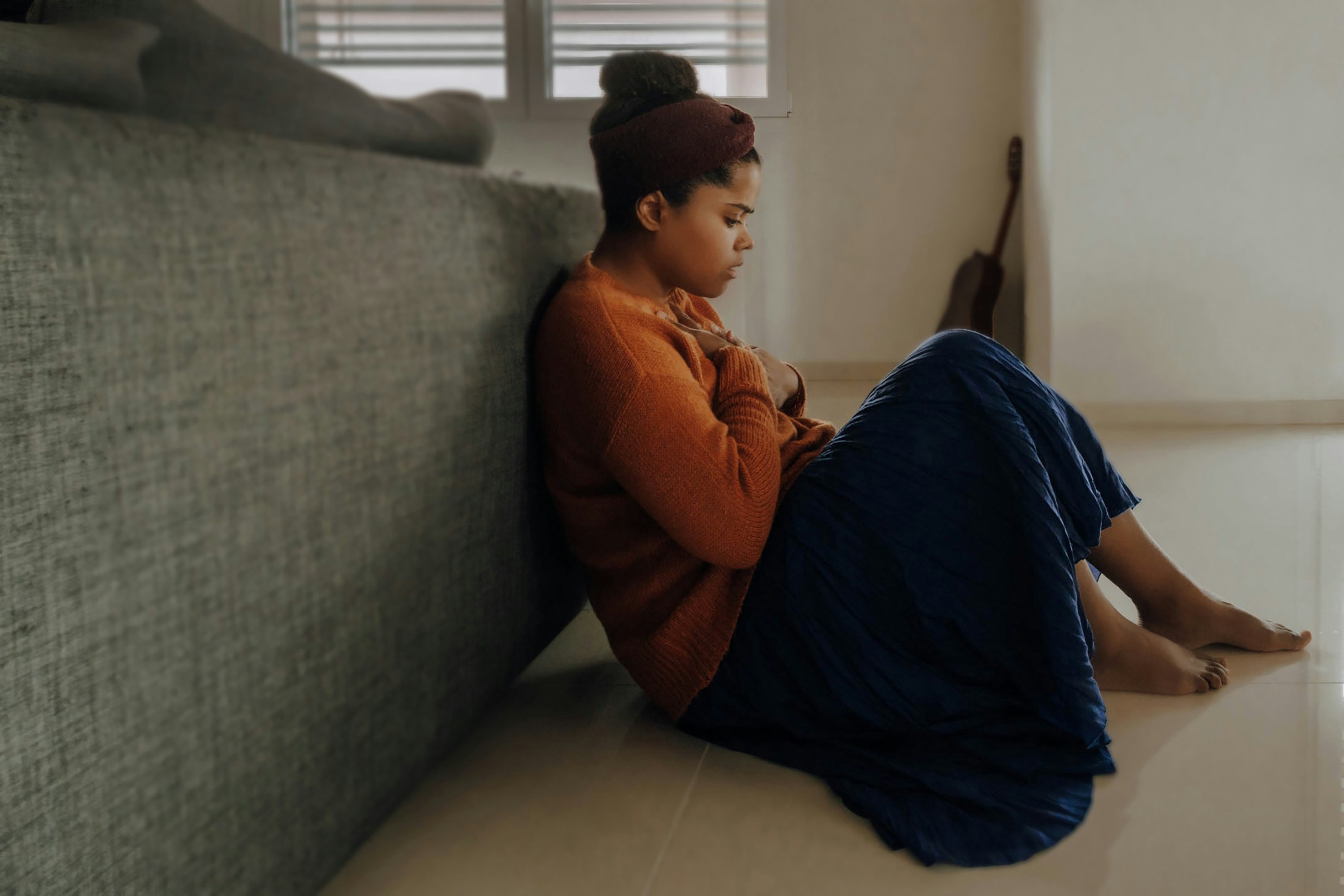 Woman sitting on the floor beside a sofa with her knees drawn up, looking down with a pensive expression in a softly lit room.