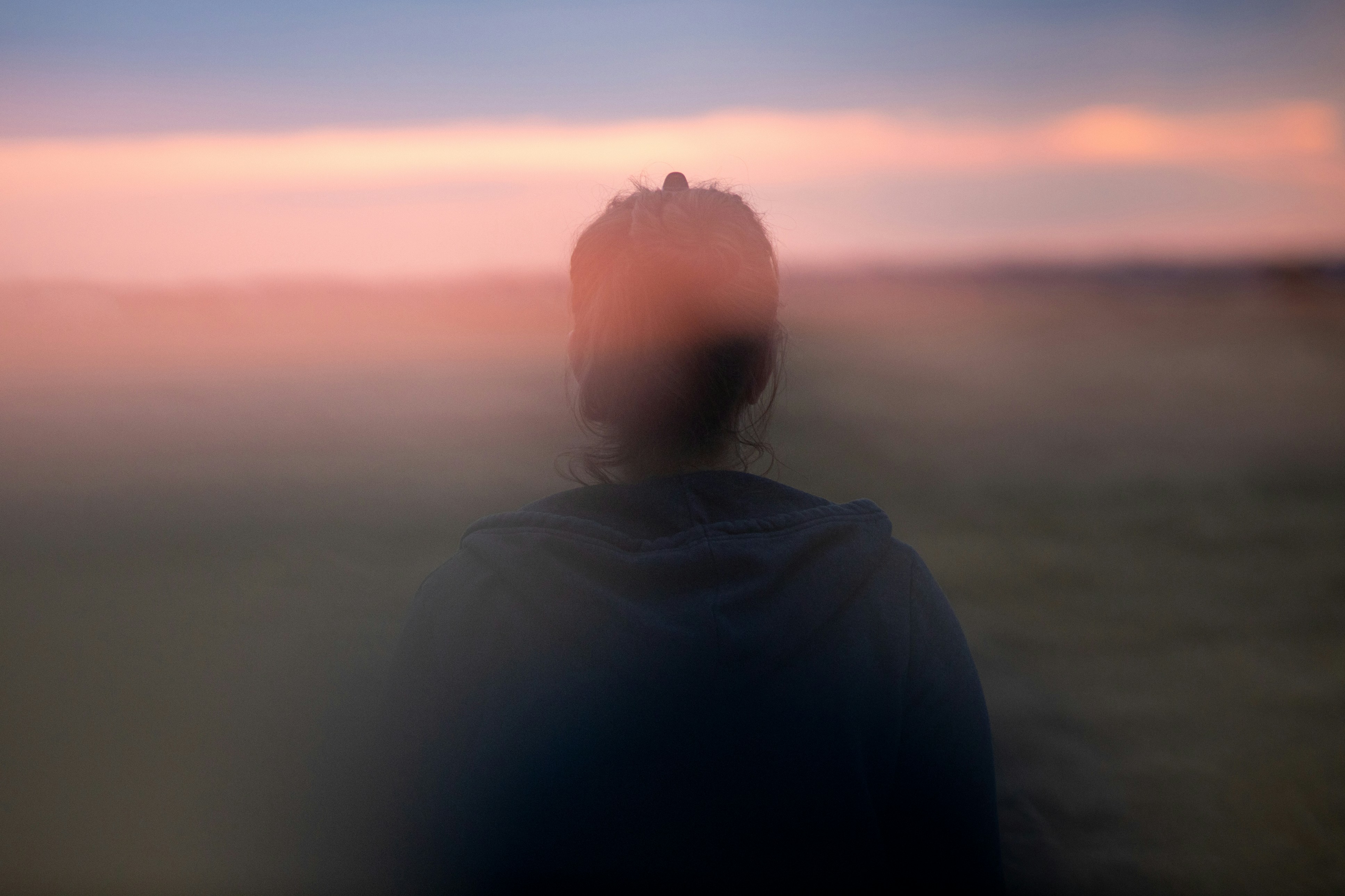 Person seen from behind standing outdoors at dusk, looking toward a softly blurred pink and blue horizon with a dreamy, contemplative atmosphere. Experiencing brain fog. 