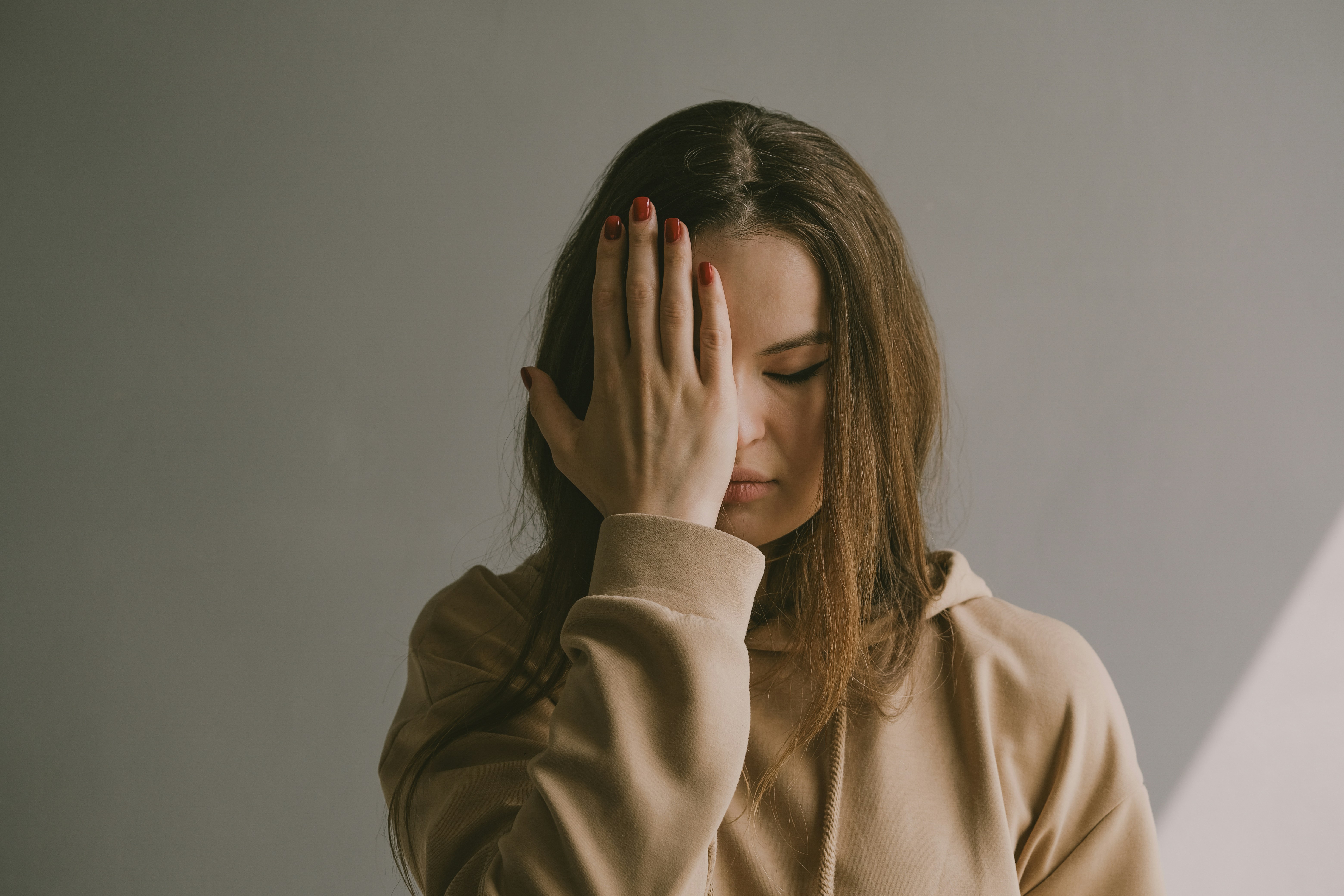 Woman in a beige hoodie covering one eye with her hand, standing against a neutral background in soft natural light.