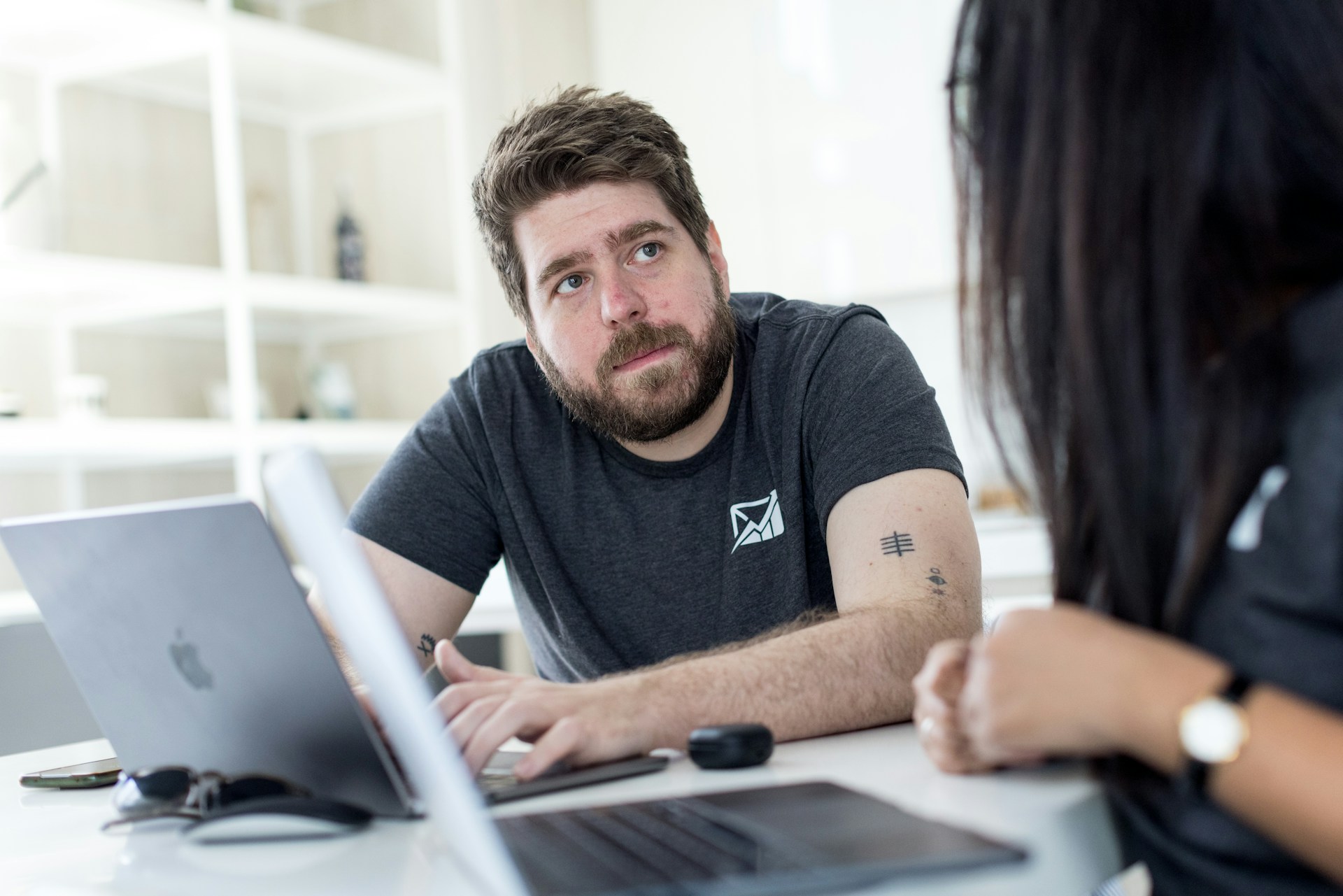 Two coworkers having a conversation at a desk with open laptops in a bright modern office.