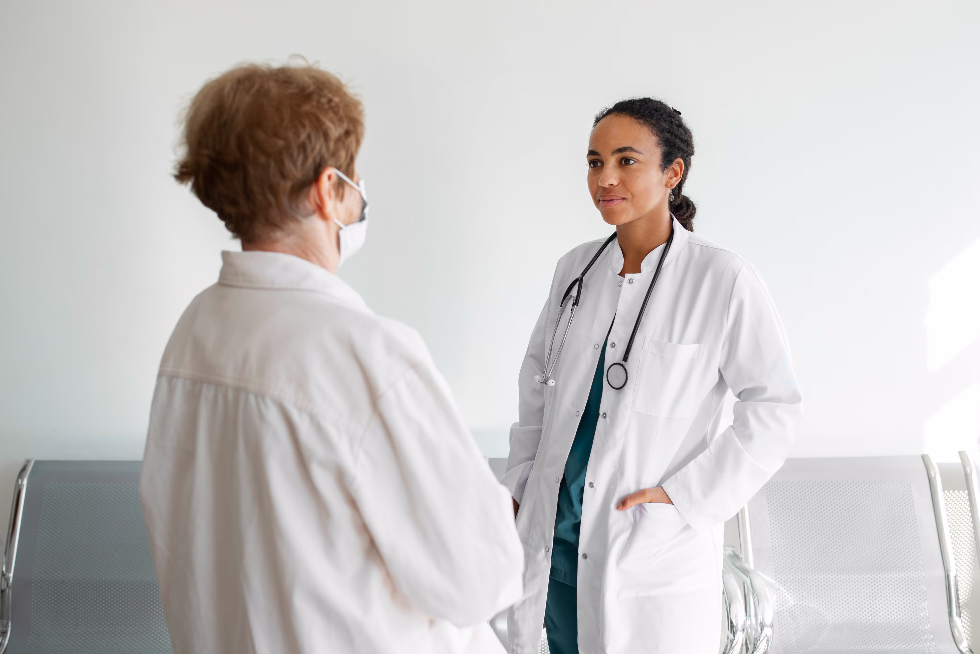 Female doctor with stethoscope speaking to older patient wearing a face mask in a medical waiting room.