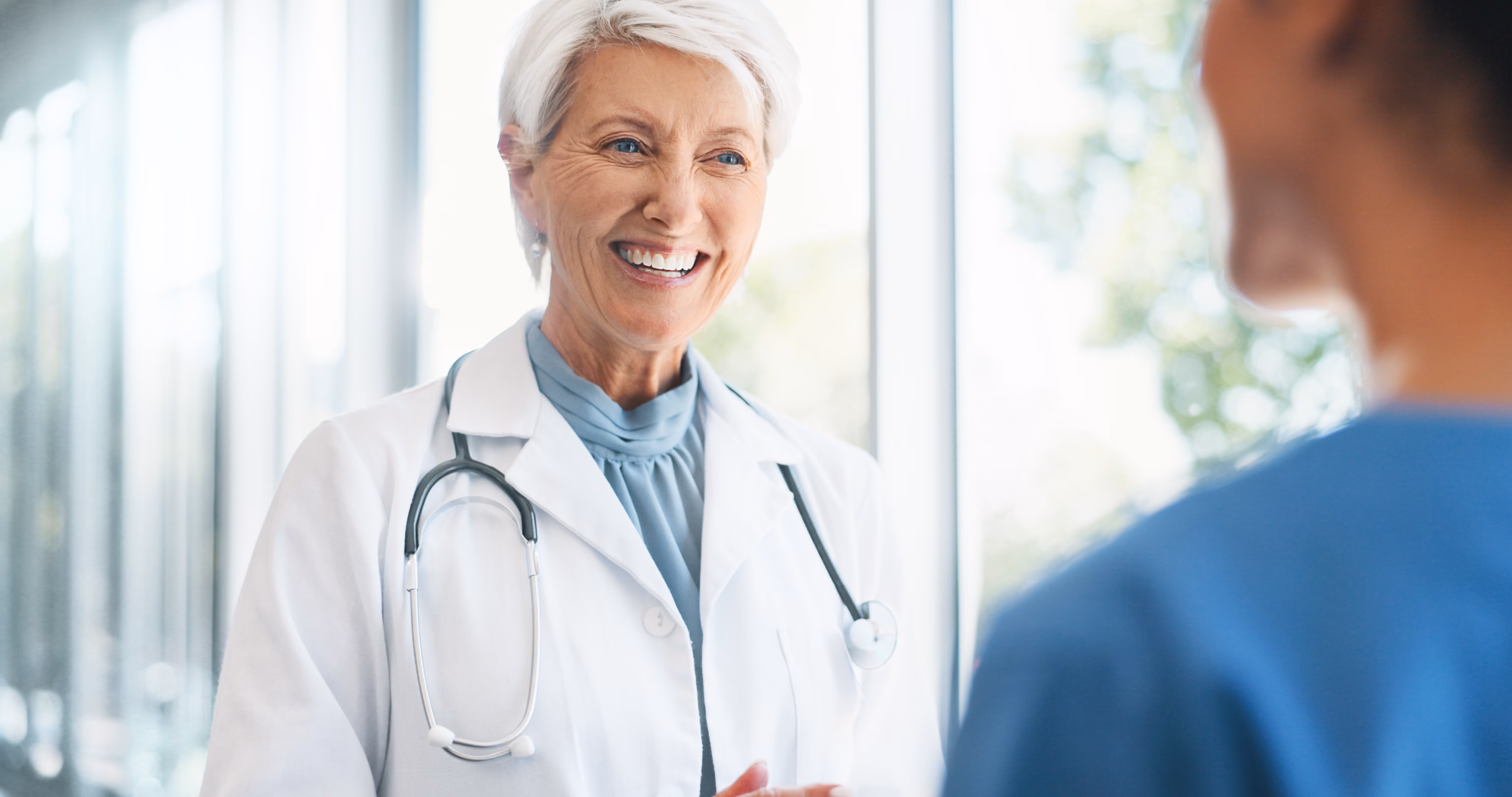 Smiling physician in a white coat and stethoscope talking with a colleague in blue scrubs in a bright clinic hallway