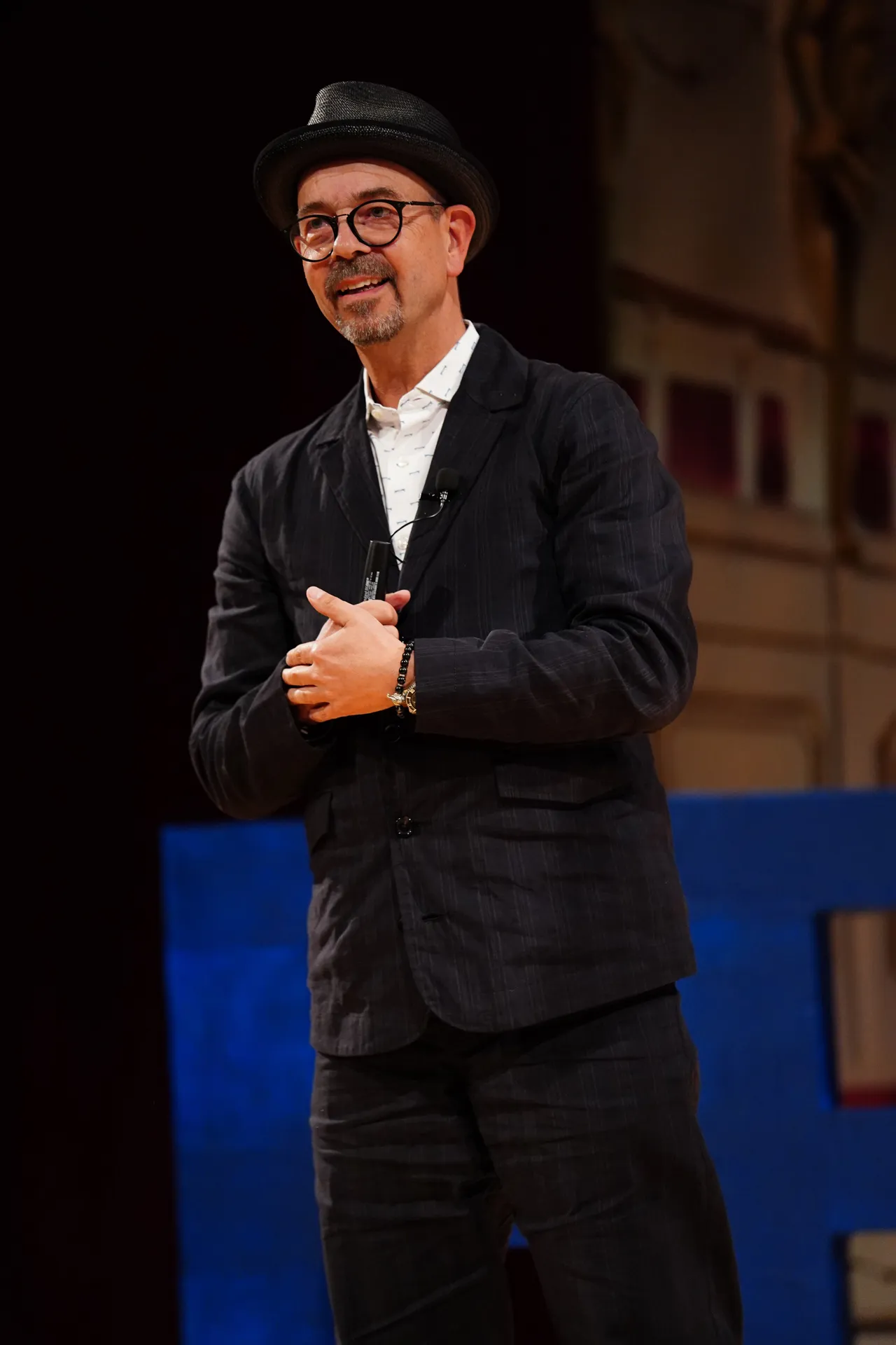 Mann mit Brille und Hut, der auf einer Bühne mit Mikrofon in der Hand spricht.