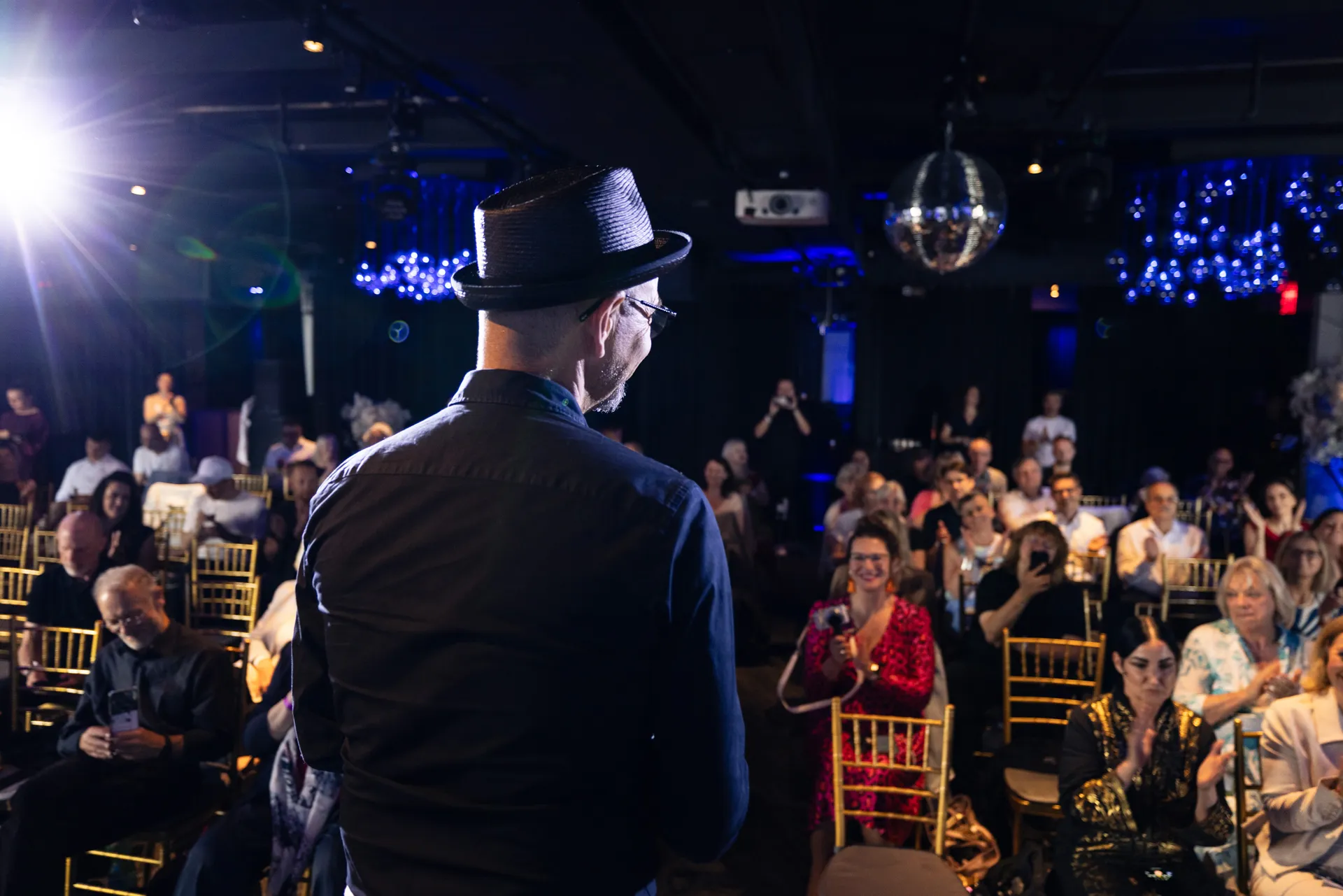 Ein Mann mit Hut und Brille spricht vor einem applaudierenden Publikum in einem dunklen Veranstaltungsraum mit blauer Beleuchtung.