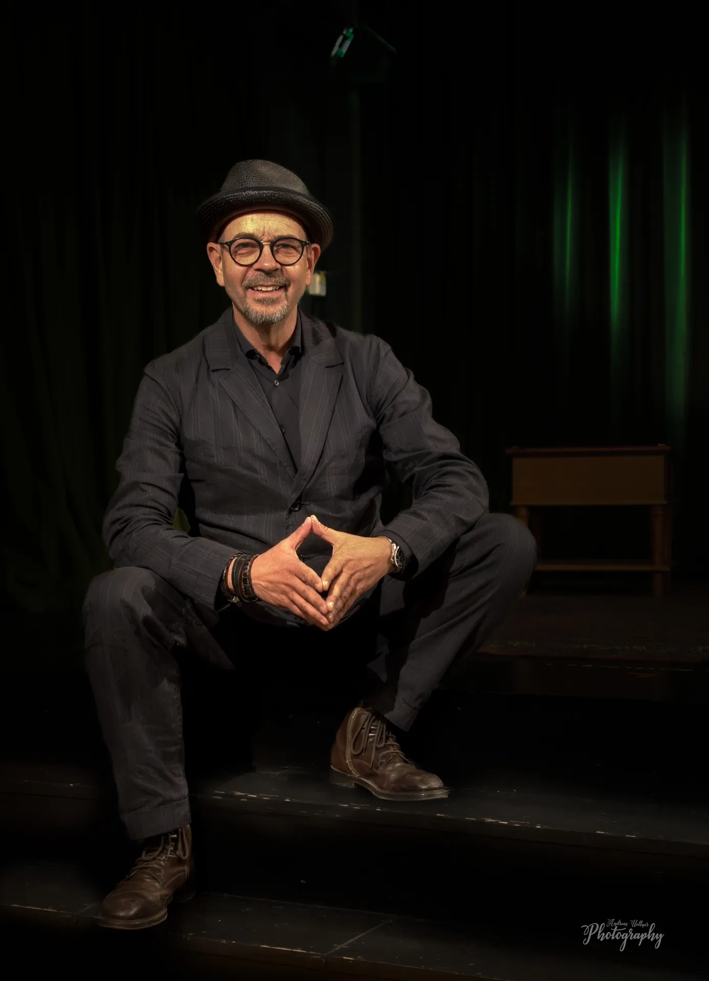 Lächelnder Mann mit Brille und grauem Hut lehnt vor reflektierender Glaswand.