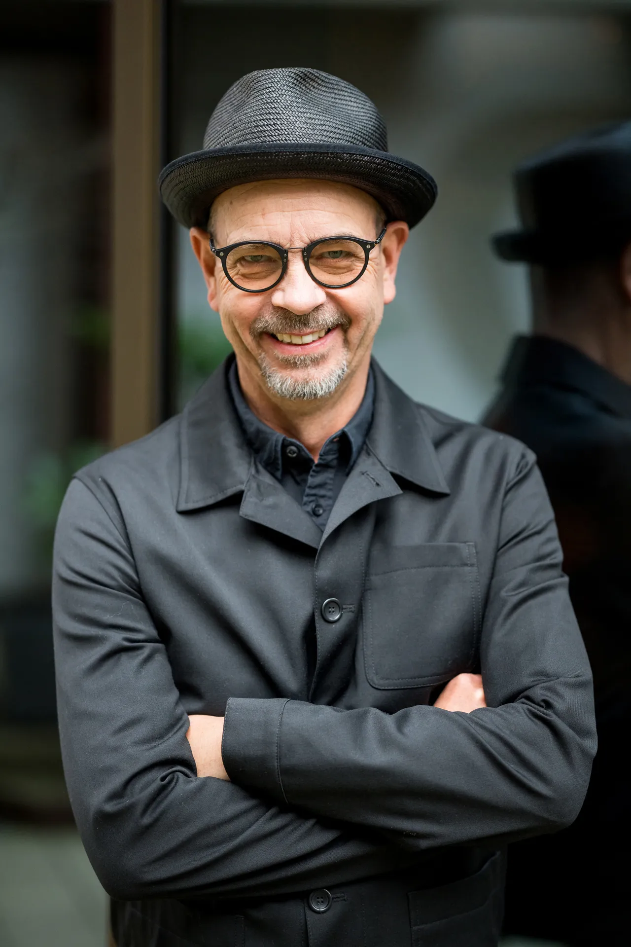 Älterer Mann mit schwarzem Hut, Brille und schwarzem Mantel lächelt mit verschränkten Armen.