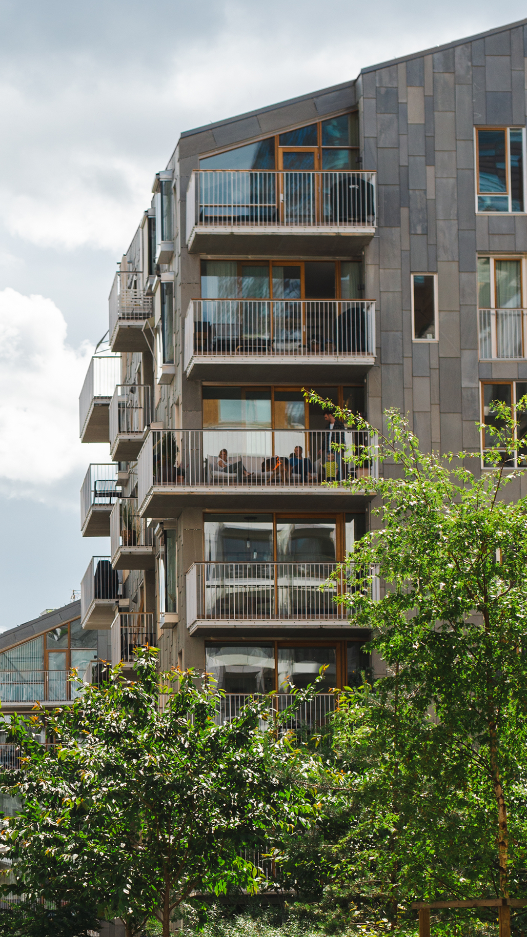 Moderne leilighetsbygning med balkonger og folk som sitter ute, omgitt av grøntområder og trær.