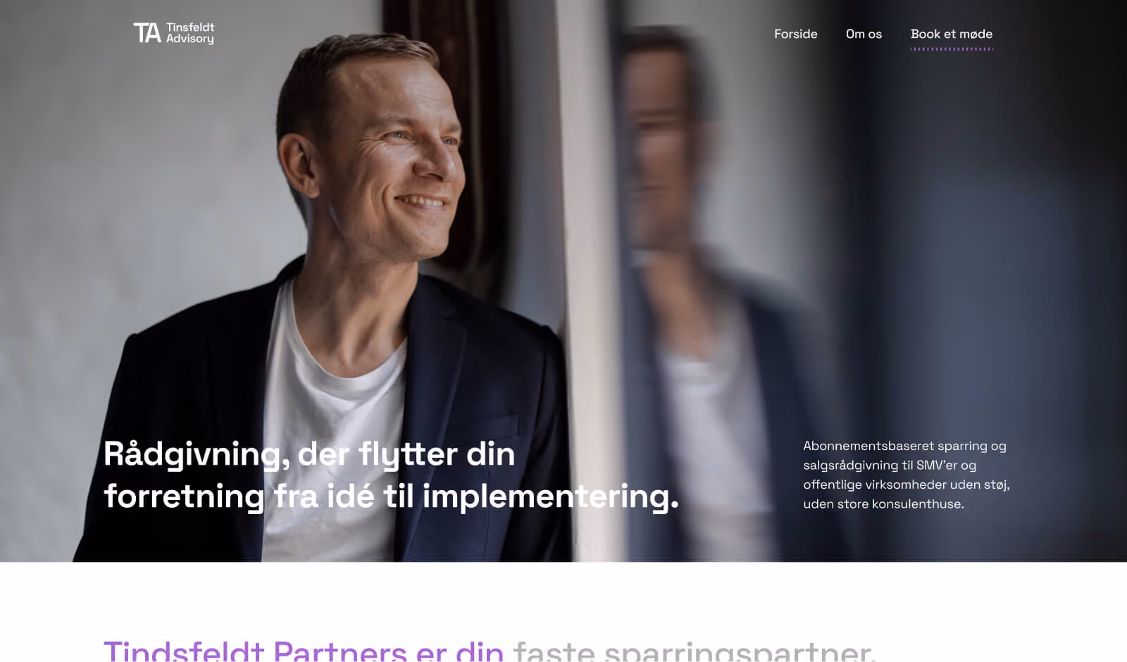Smilende mand i mørk blazer og hvid t-shirt ser ud af vindue med sin refleksion ved siden af sig.