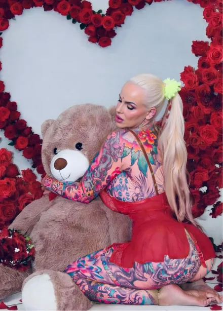Tattooed woman in a red outfit hugging a large teddy bear in front of a heart-shaped rose decoration.